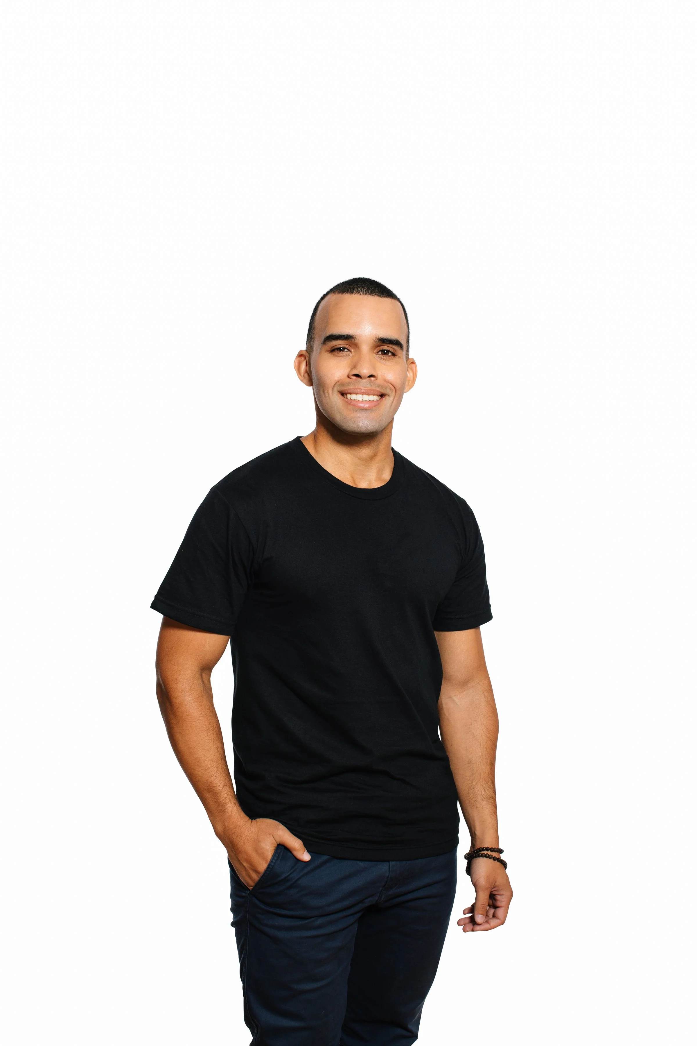 A man with short dark hair, wearing a black t-shirt and dark pants, smiling, with his right hand in his pocket and standing against a plain white background.