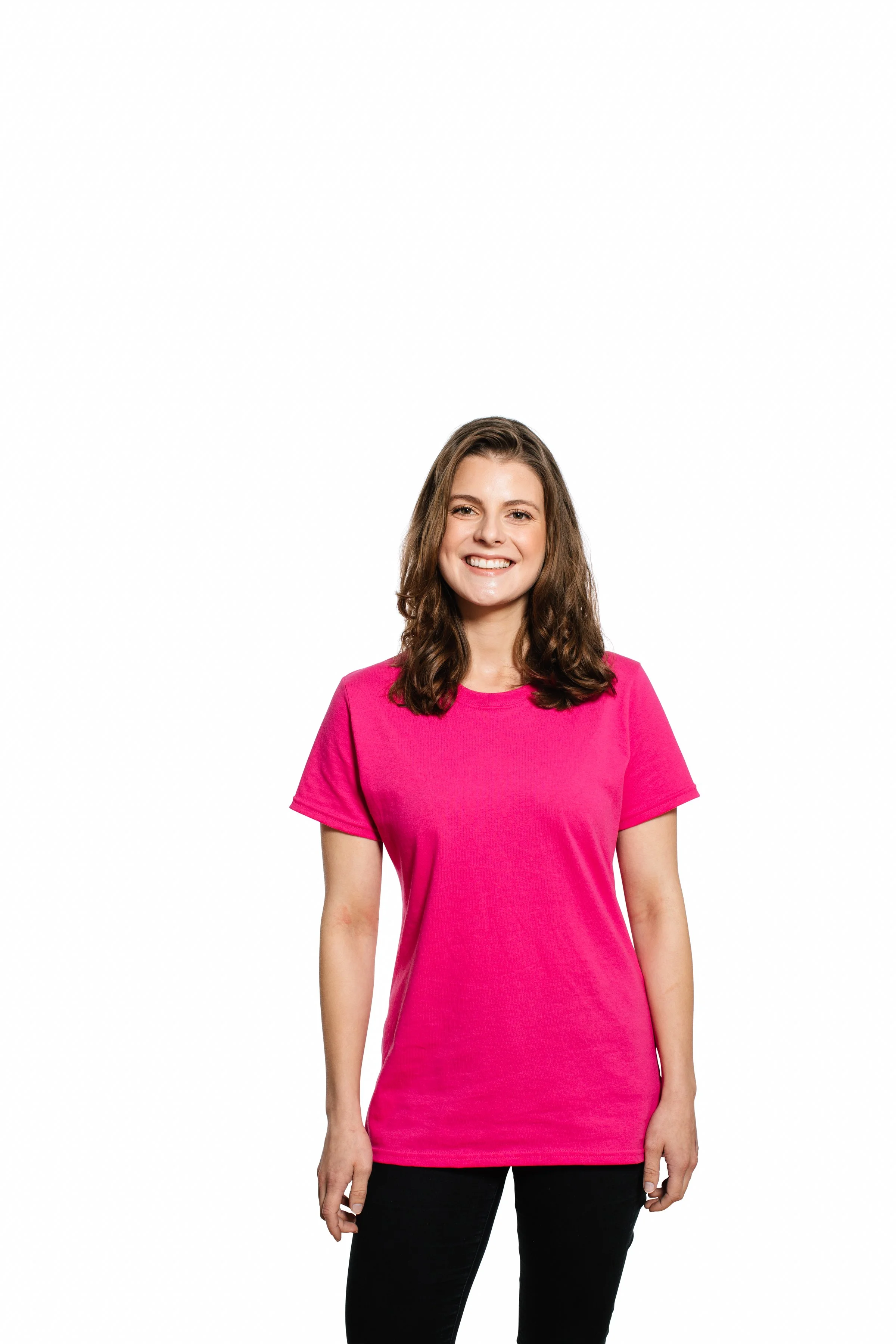 A young woman with long brown hair smiling, wearing a bright pink t-shirt and black pants, standing against a plain white background.