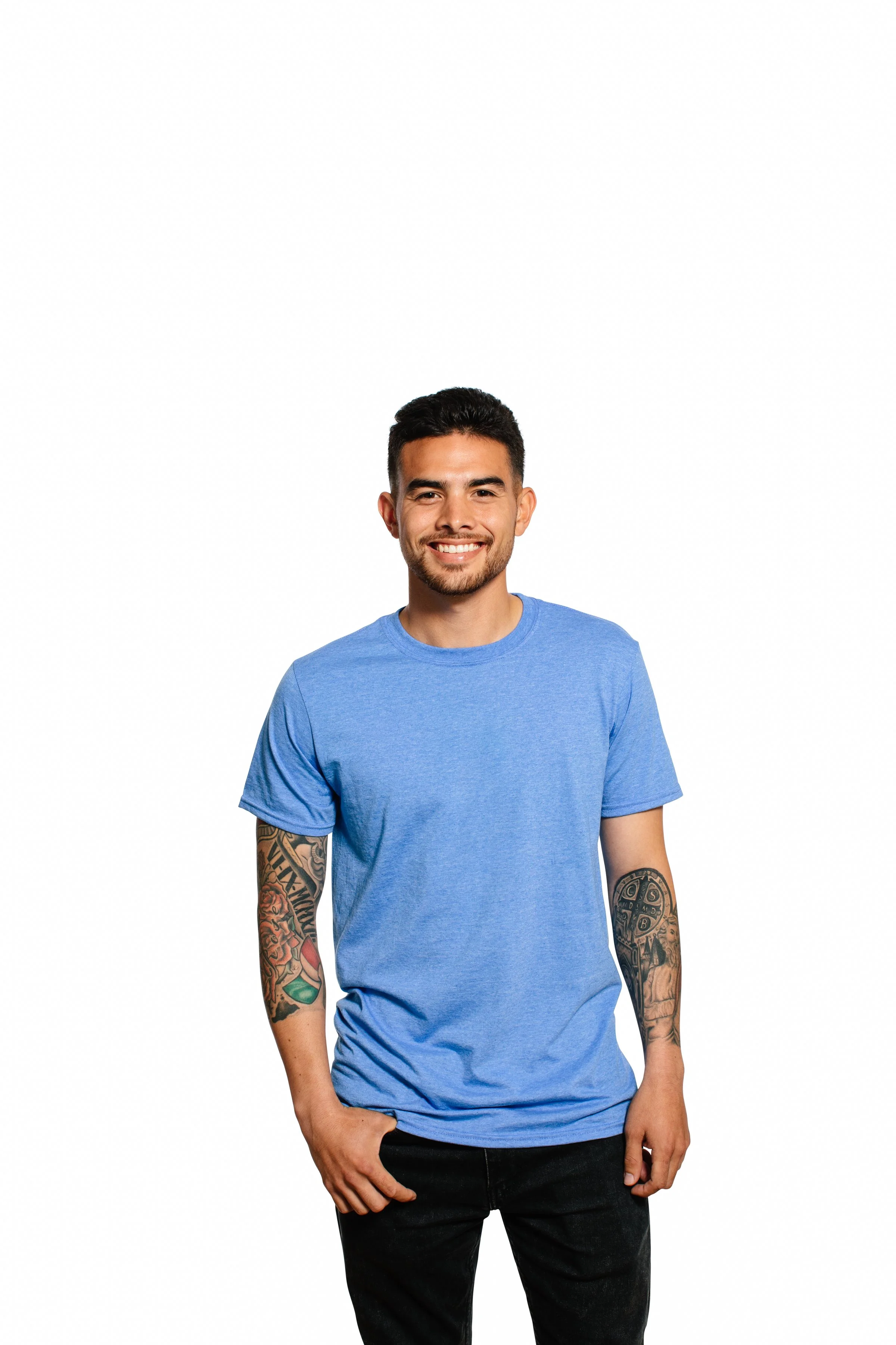 A smiling young man with tattoos on his arms wearing a blue t-shirt and black jeans, standing against a plain white background.