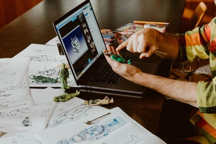 Person working on a laptop with toy soldiers and sketches on a table.