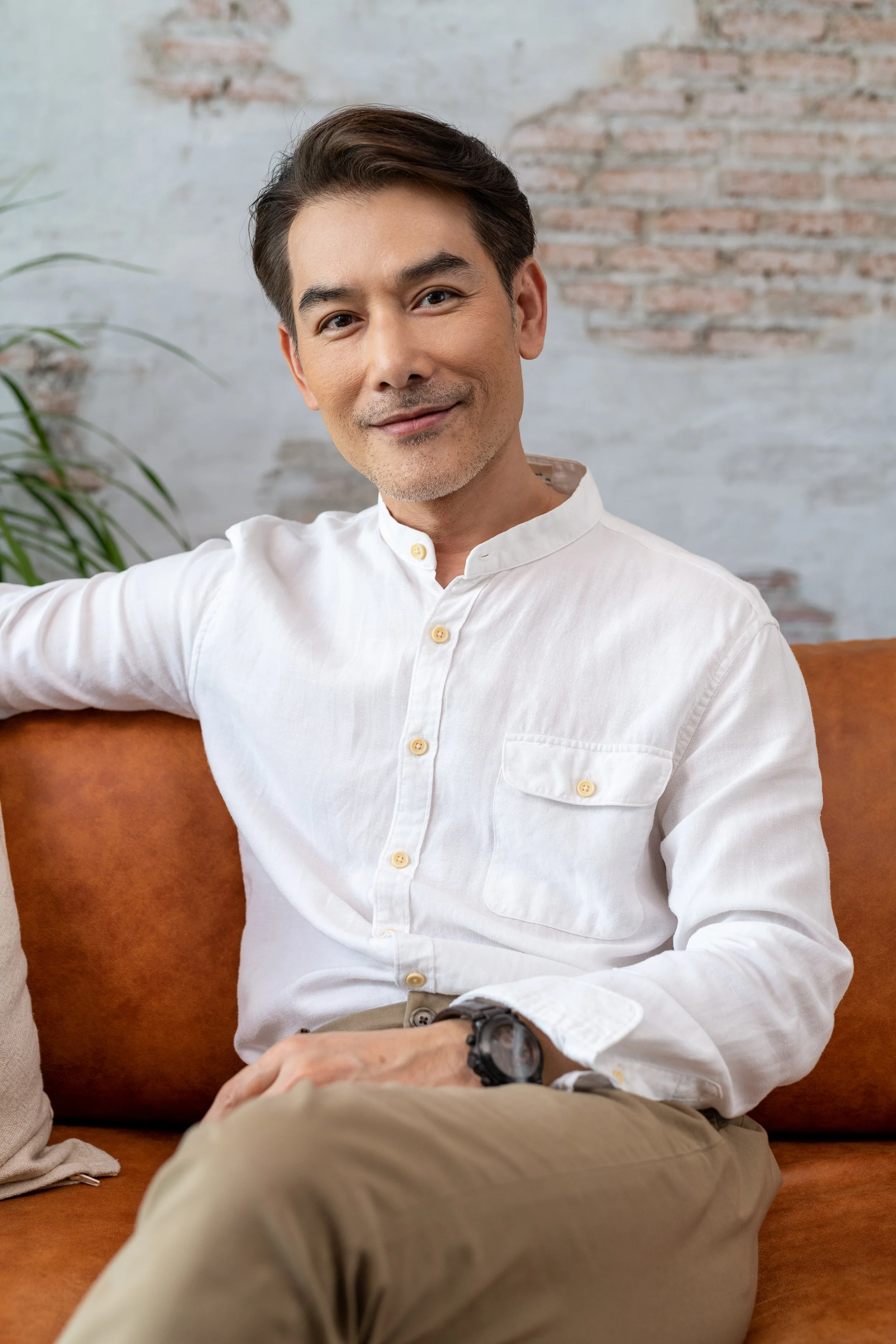 A man with dark hair wearing a white button-up shirt and beige pants sitting on a brown sofa with a brick wall and a plant in the background.