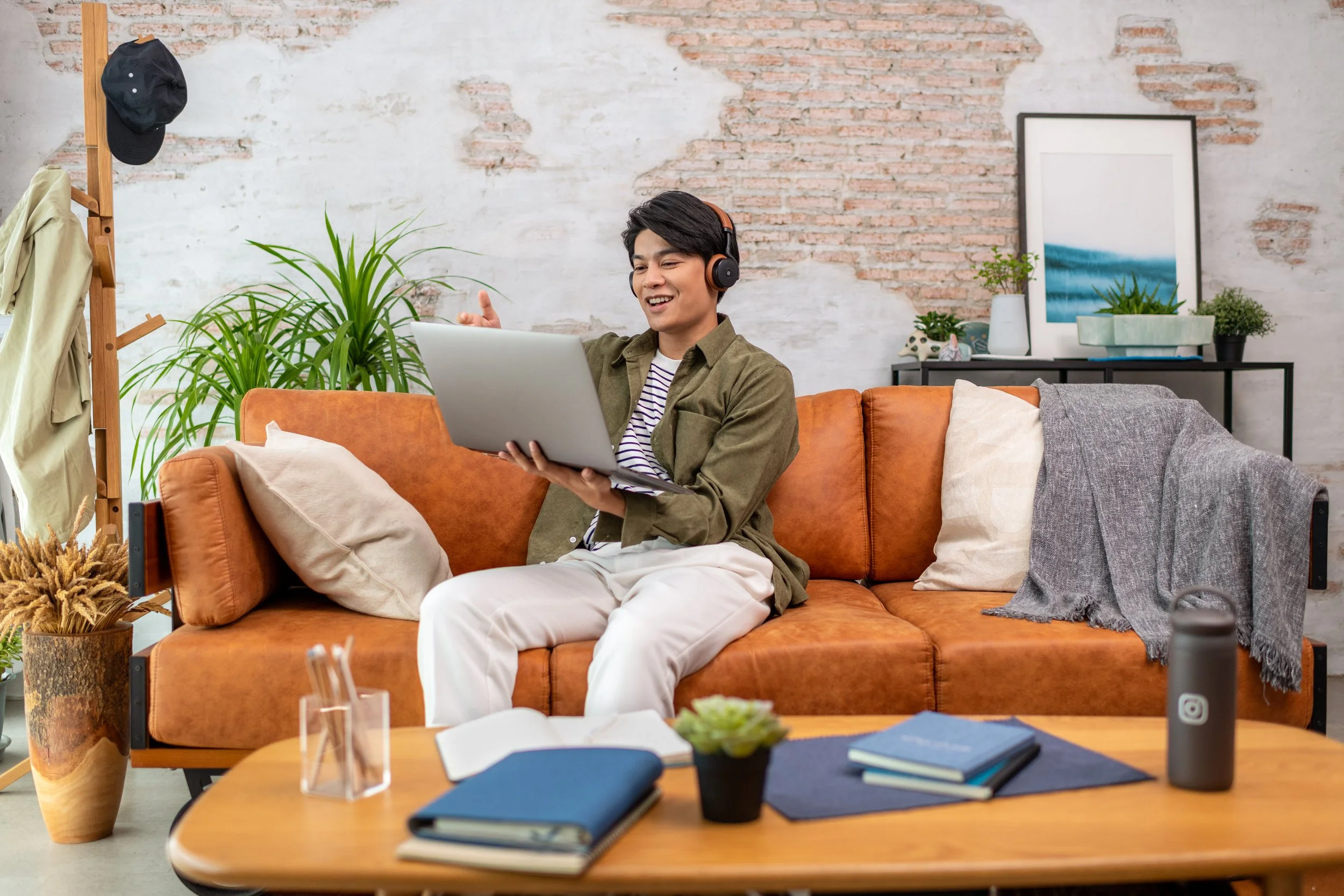 A young man sitting on an orange couch with beige cushions, wearing headphones, holding a laptop, smiling and giving a thumbs-up gesture. The room has a brick wall, plants, framed art, and a wooden coat rack with clothing.