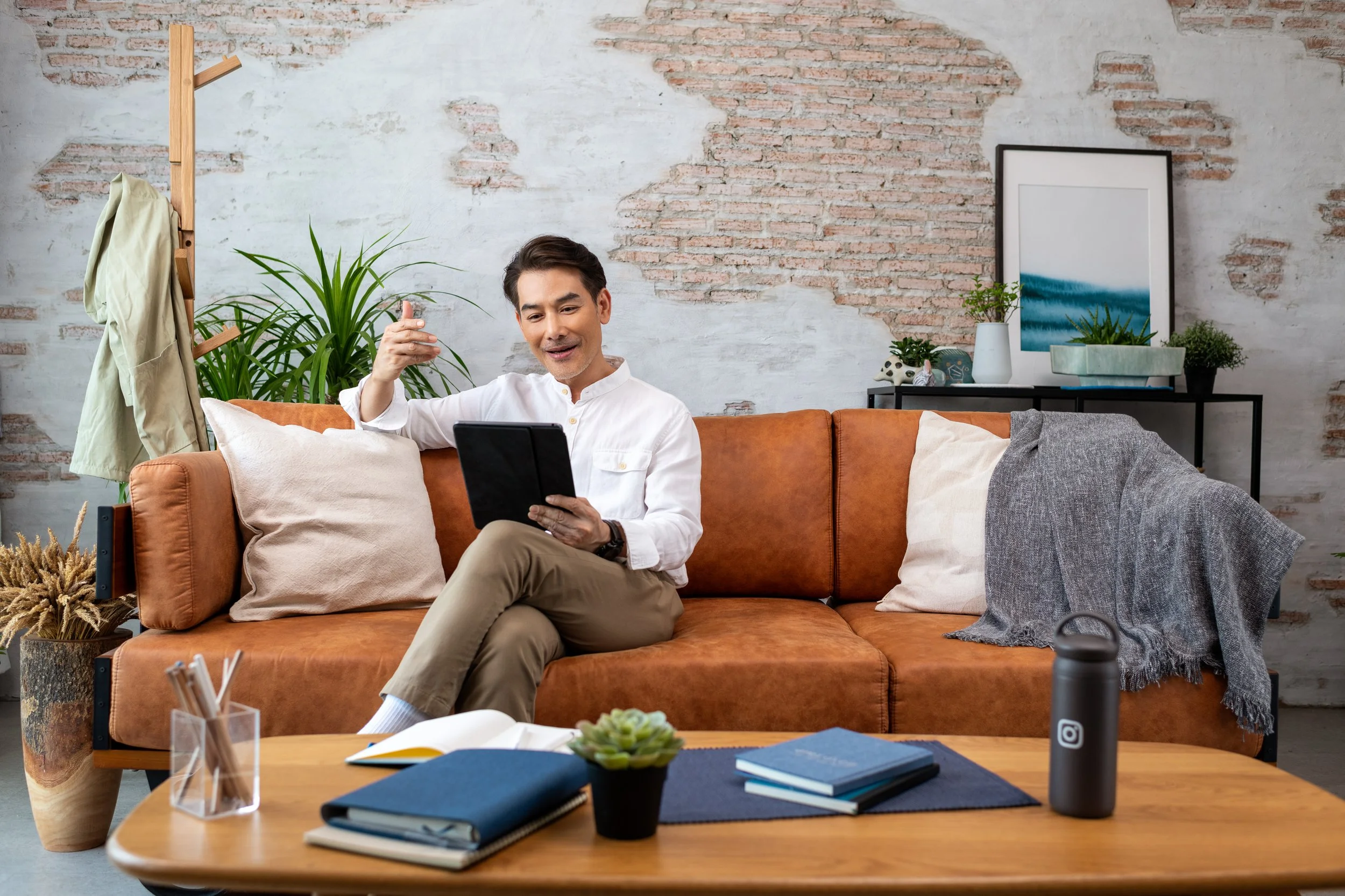 A man sitting on a brown sofa in a living room, holding a tablet and smiling. The room has a rustic brick wall, a wooden coat rack with a jacket, plants, framed artwork, and a coffee table with notebooks, a succulent, and a black water bottle.