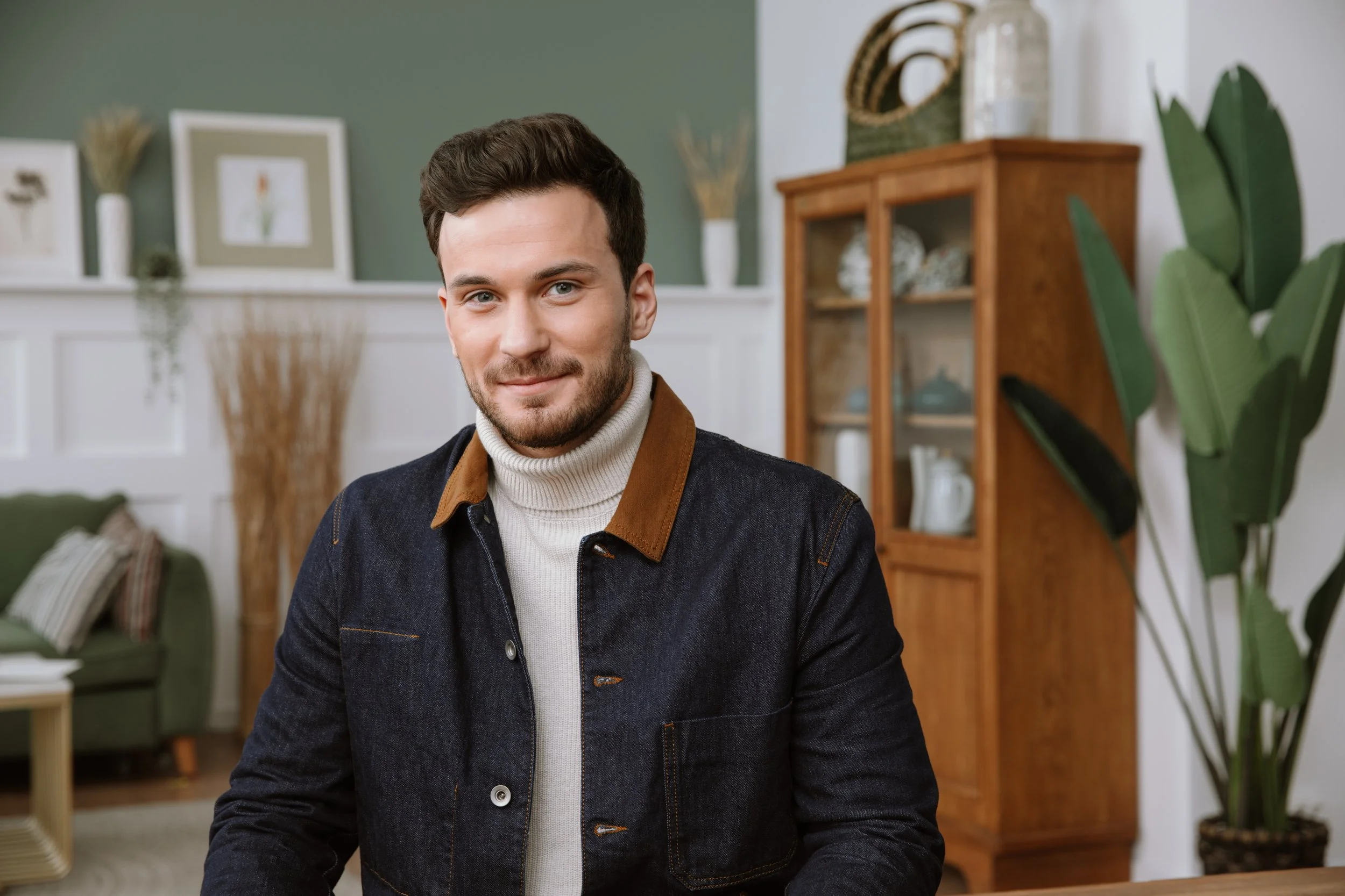 A young man with dark hair and a beard, wearing a denim jacket with a tan collar and a white turtleneck, sitting in a cozy living room with green walls, framed artwork, and houseplants.