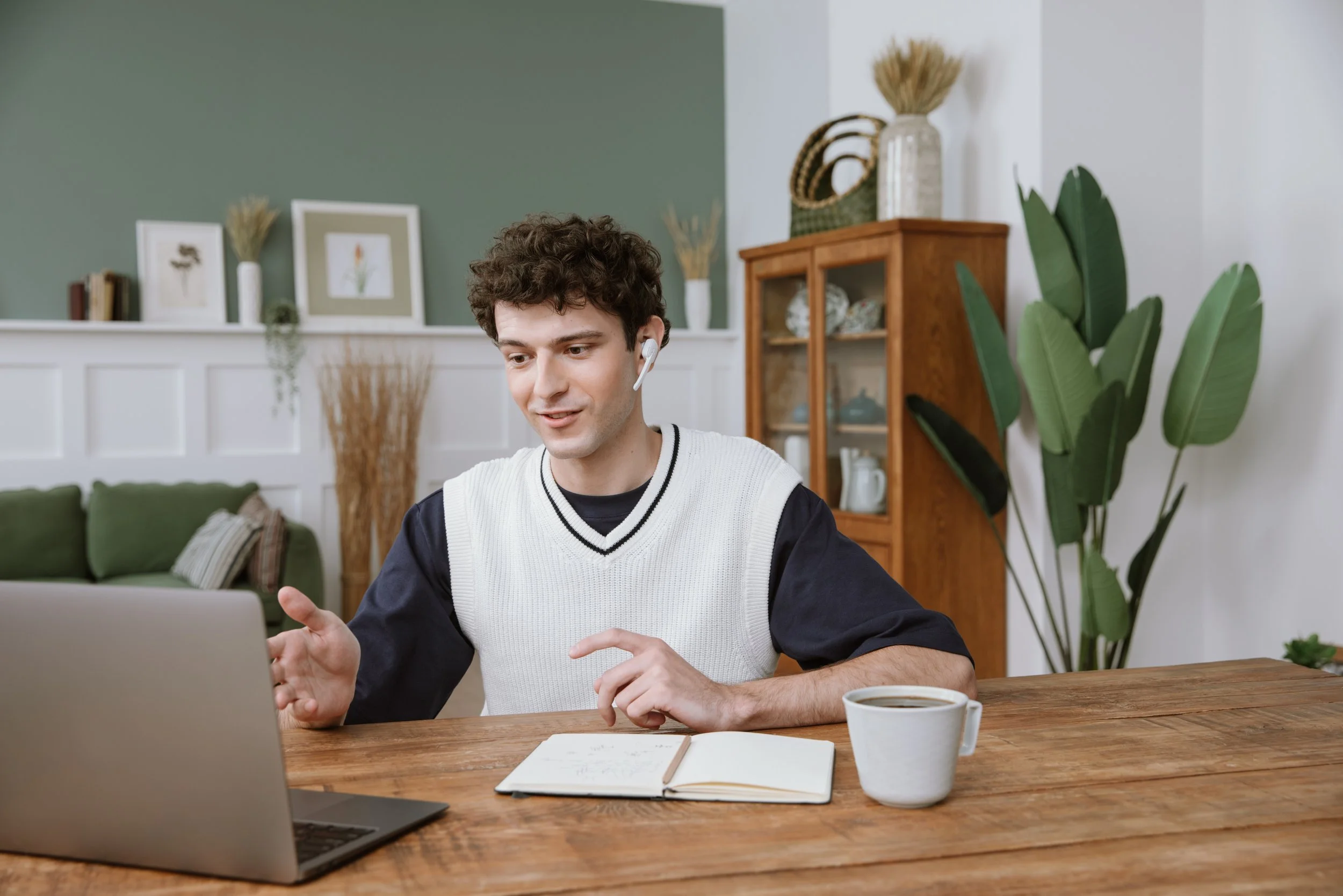 A young man with curly hair wearing earbuds, a white vest over a black shirt, sitting at a wooden table with a laptop, notebook, and cup of coffee, in a living room with plants, framed artwork, and a cabinet in the background.