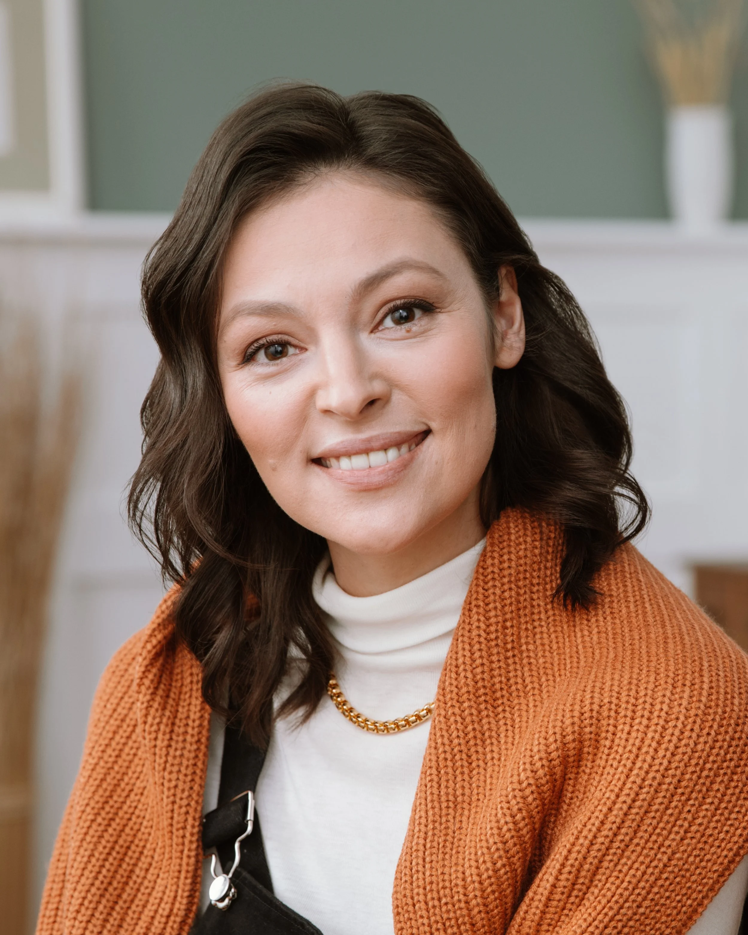 A woman with long dark brown wavy hair, smiling, wearing a white turtleneck, orange knitted cardigan, gold chain necklace, and a black bag with a strap.