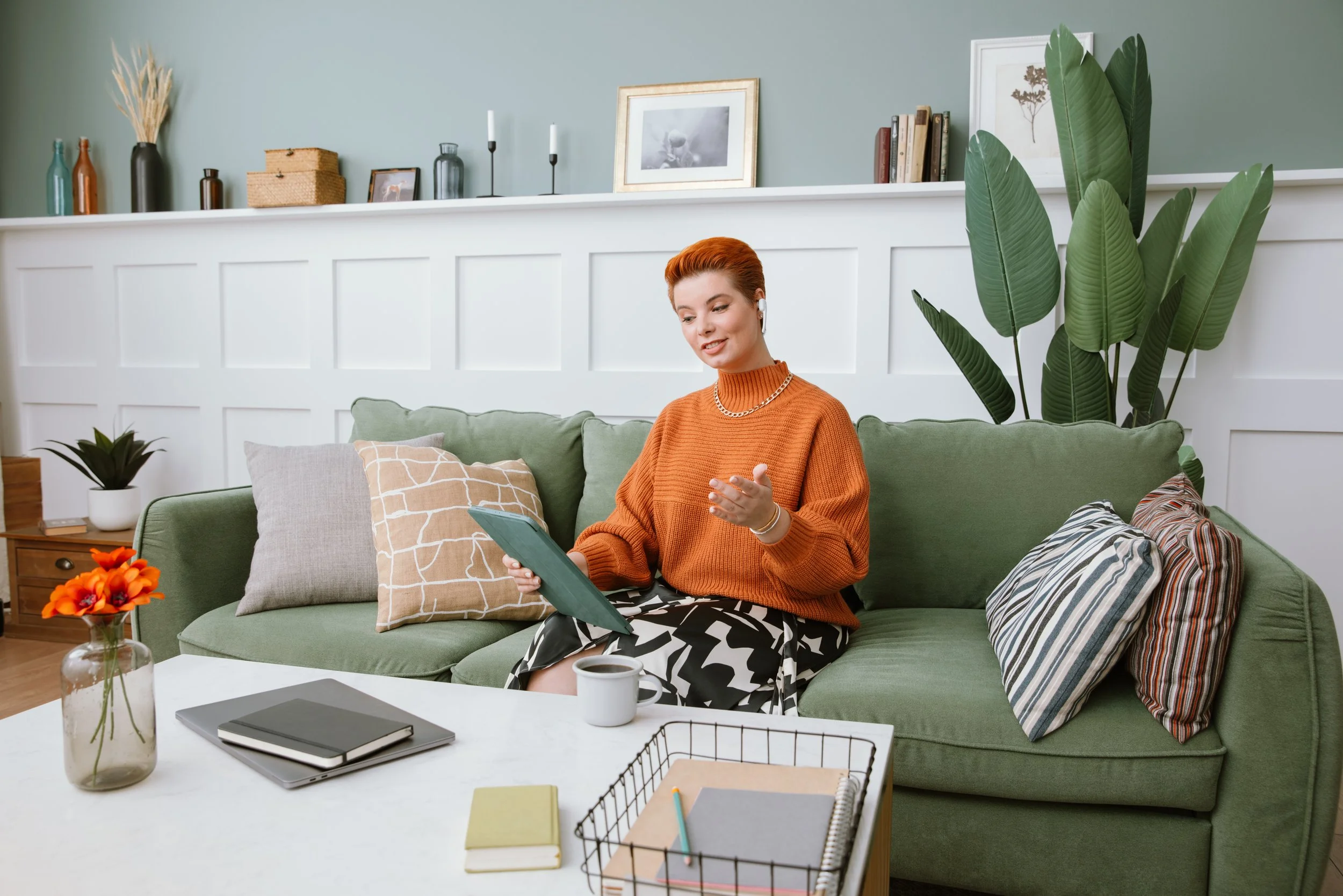 Woman with short red hair sitting on a green couch, wearing an orange sweater and a gold chain, using a tablet in a cozy living room with decorative pillows, a coffee table with books and a mug, and a large plant nearby.