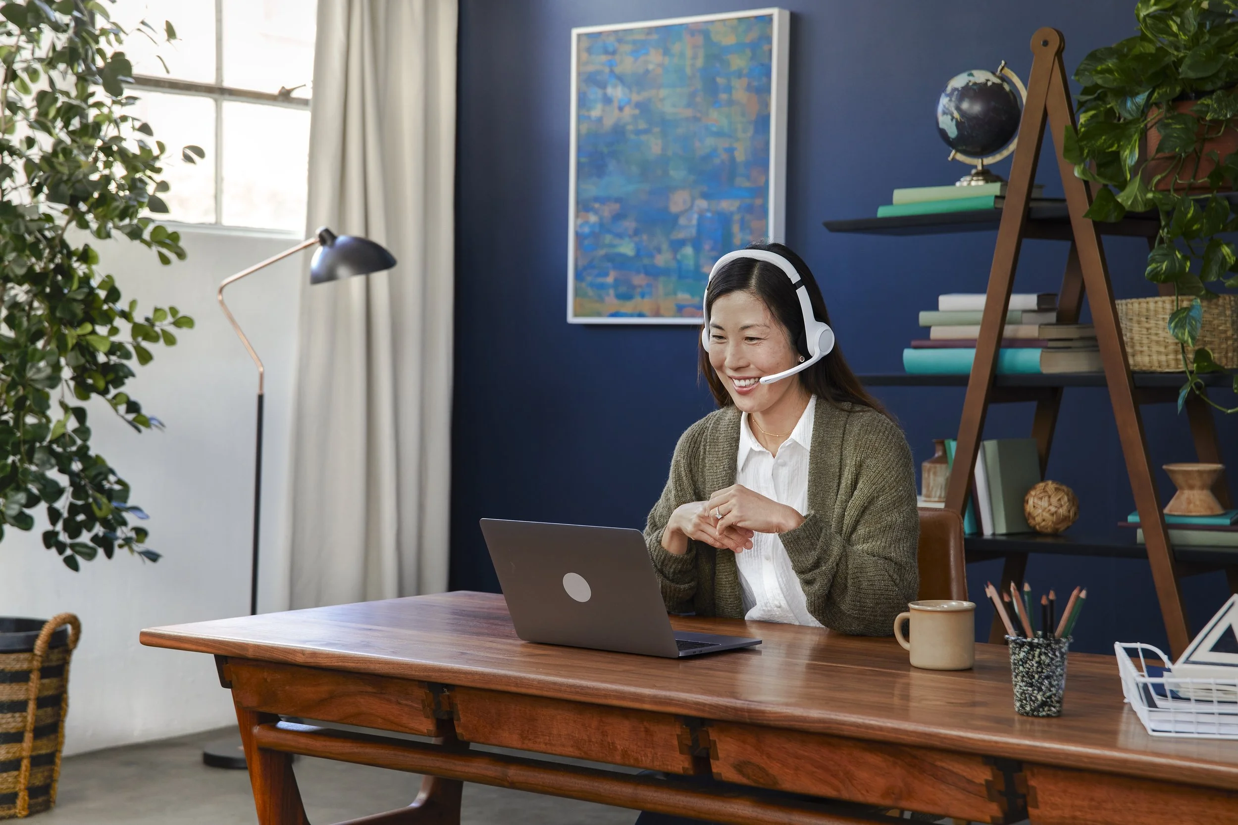 A woman in a headset smiling while working on a laptop at a wooden desk in a home office, with a navy blue wall, large window, bookshelf, and plants in the background.