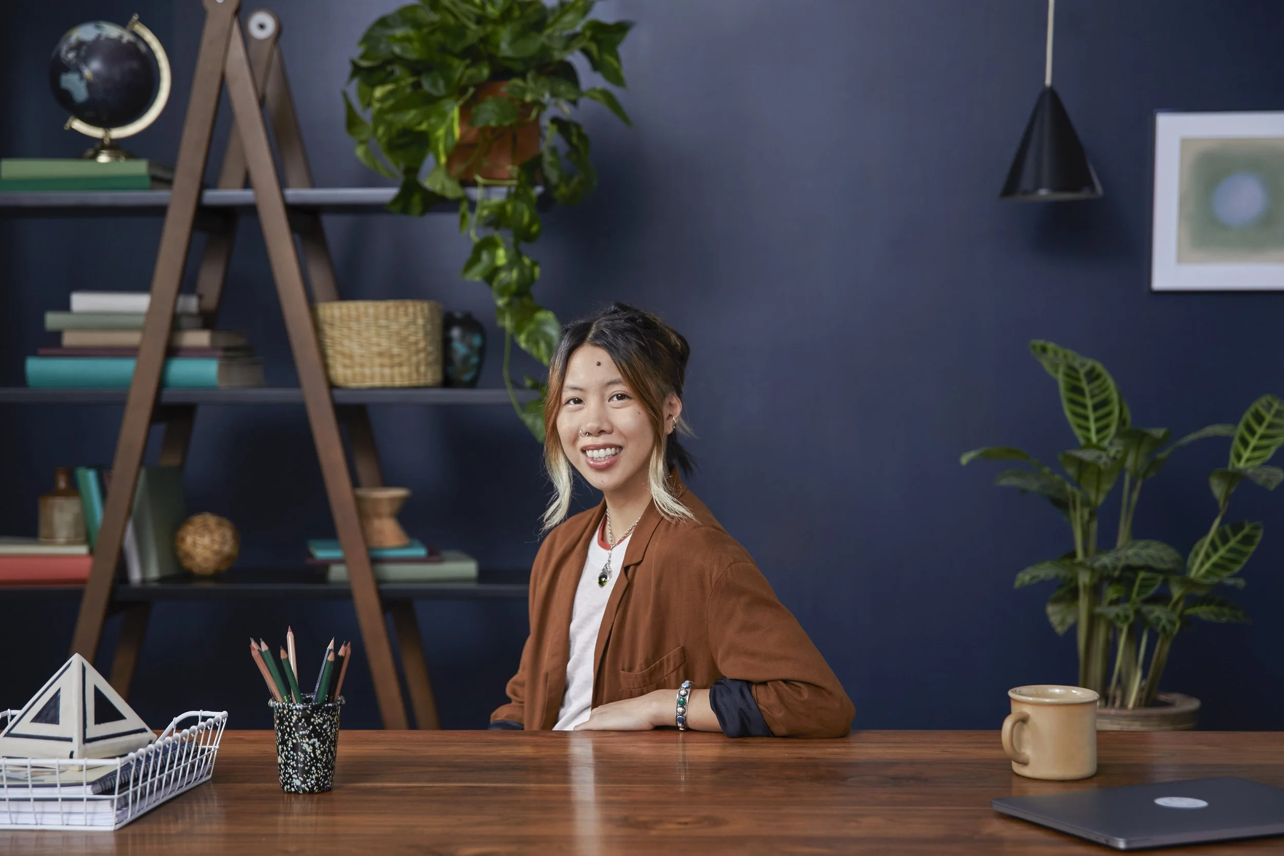A young woman with dark hair and light-colored highlights, smiling while sitting at a wooden desk in a room with dark blue walls and green plants. There are various objects on the desk, including a black and white patterned cup with colored pencils, 