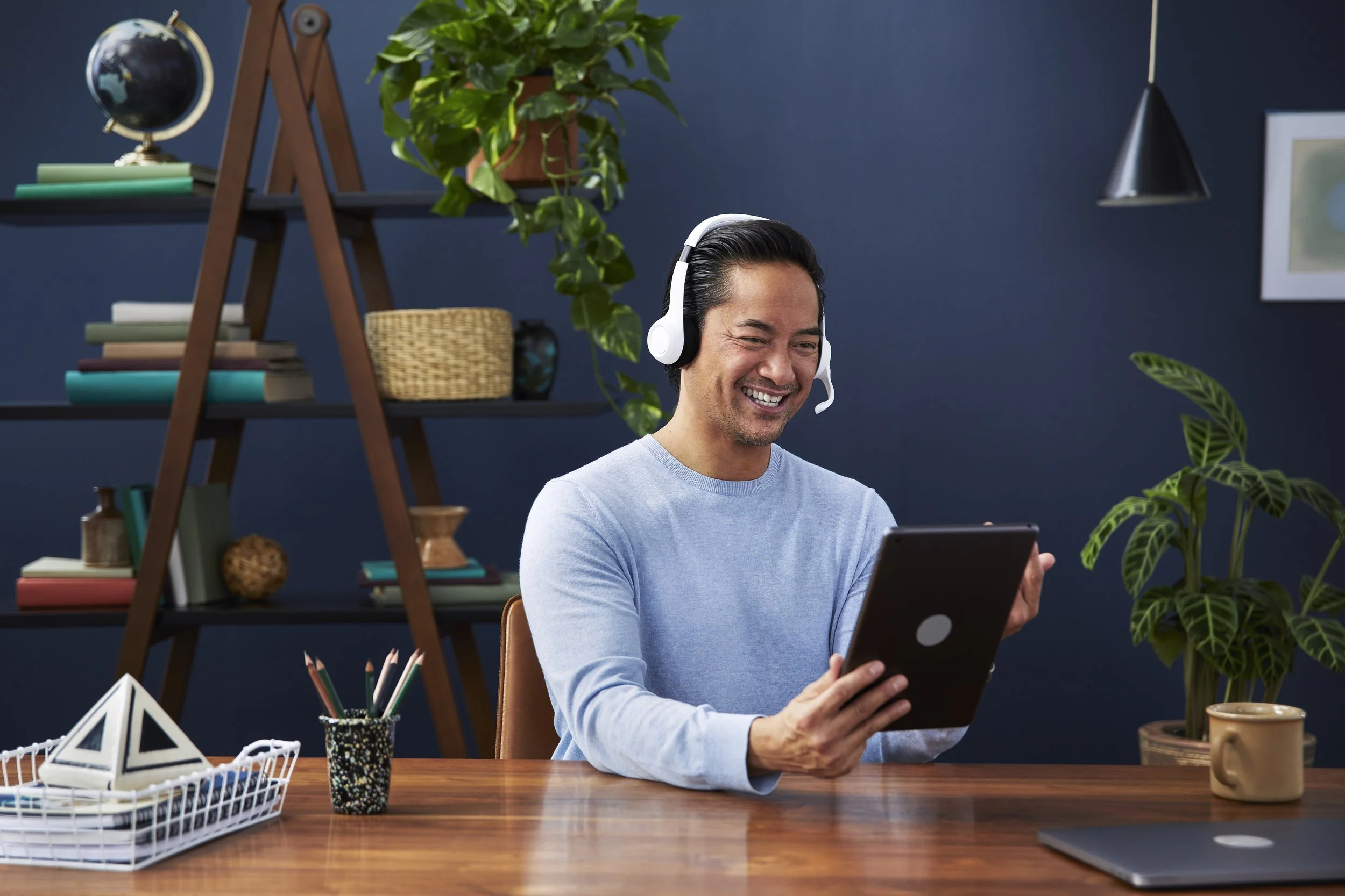 A man wearing a light blue sweater and a headset is sitting at a wooden table, smiling and looking at a tablet. The background has a blue wall with shelves filled with books, a globe, and plants. There are decorative items on the table, including a g