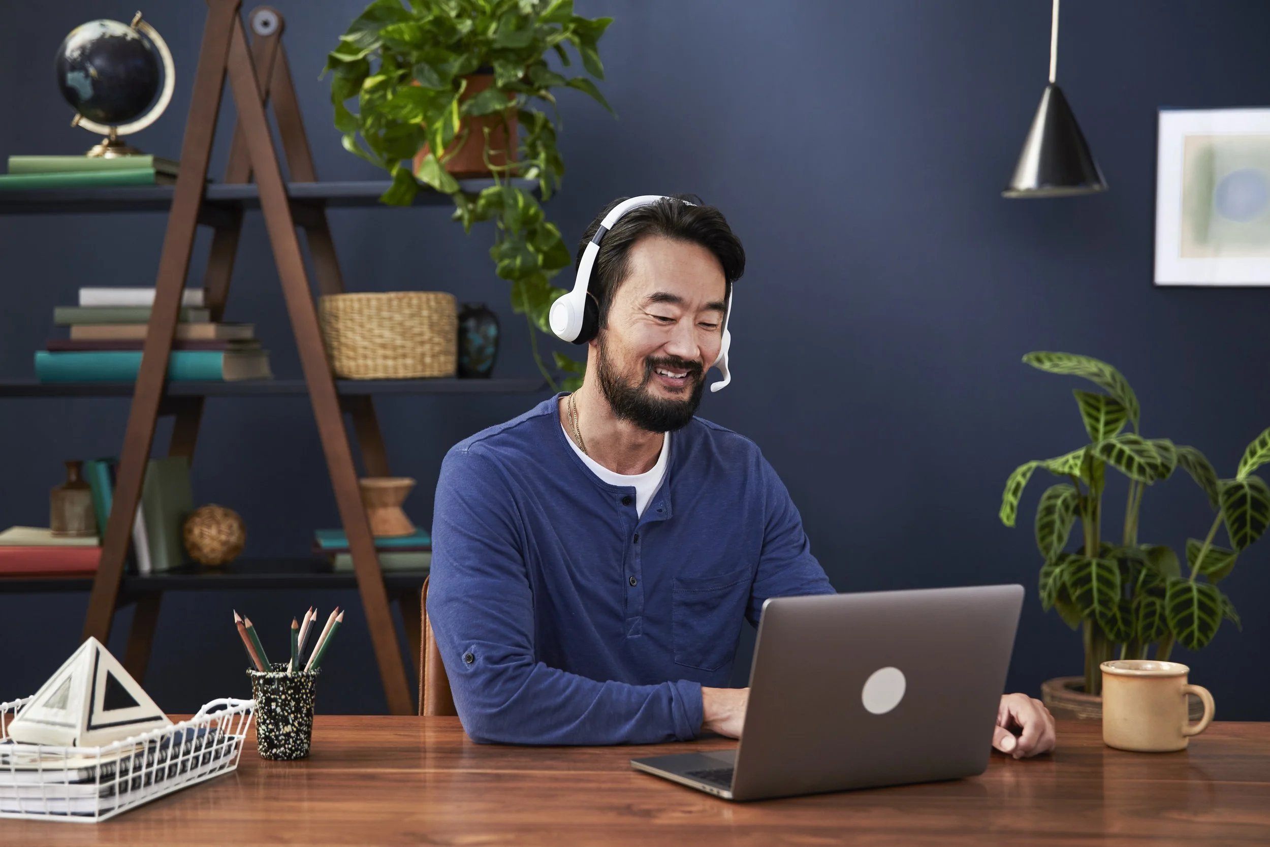 A man with a dark beard and dark hair, wearing a blue shirt, sitting at a wooden desk with a laptop in front of him. He is smiling and wearing white headphones. The background includes a blue wall with a bookshelf, some plants, and framed artworks.