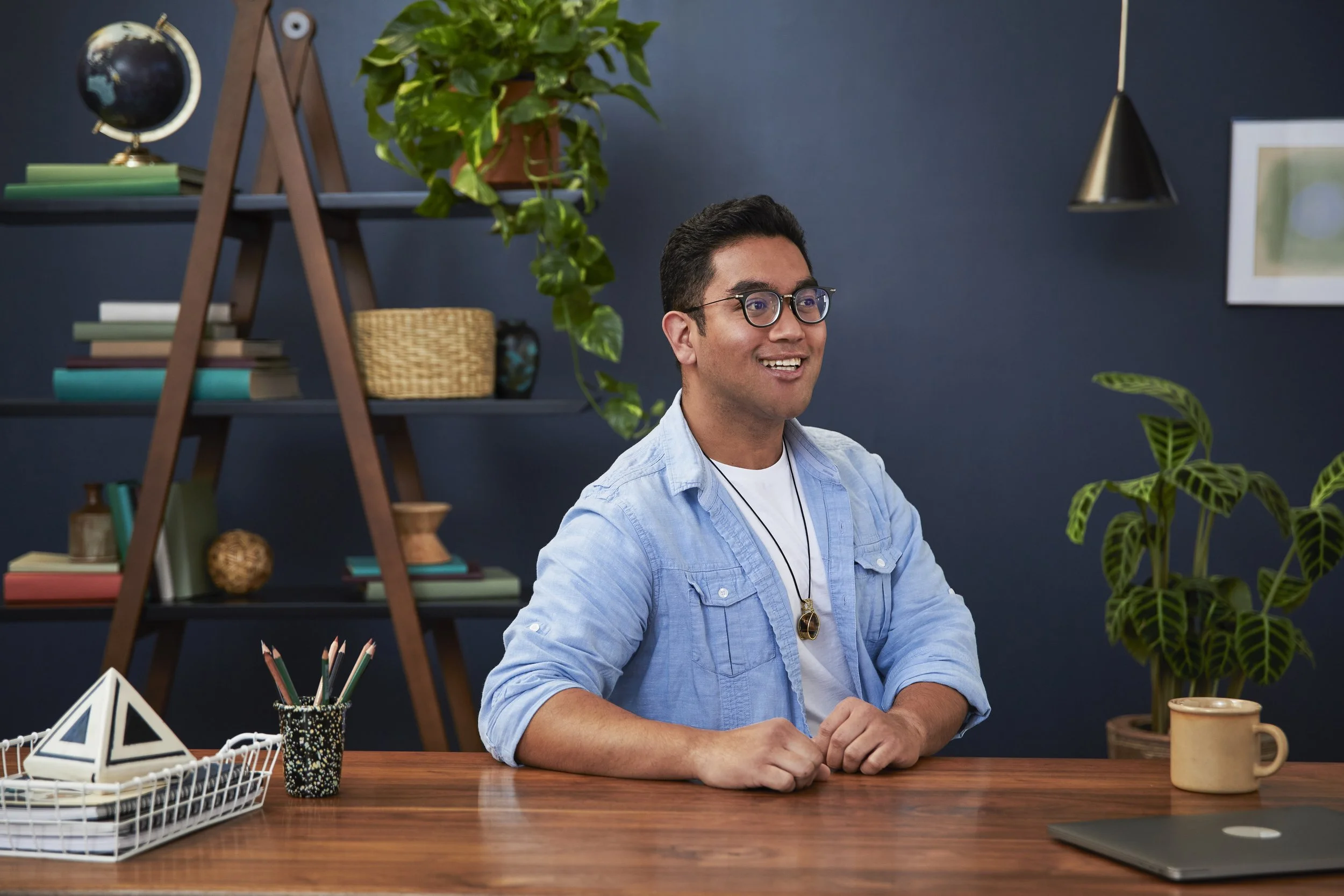 A young man smiling and sitting at a desk in a workspace decorated with plants, books, and office decor, with a dark wall background.