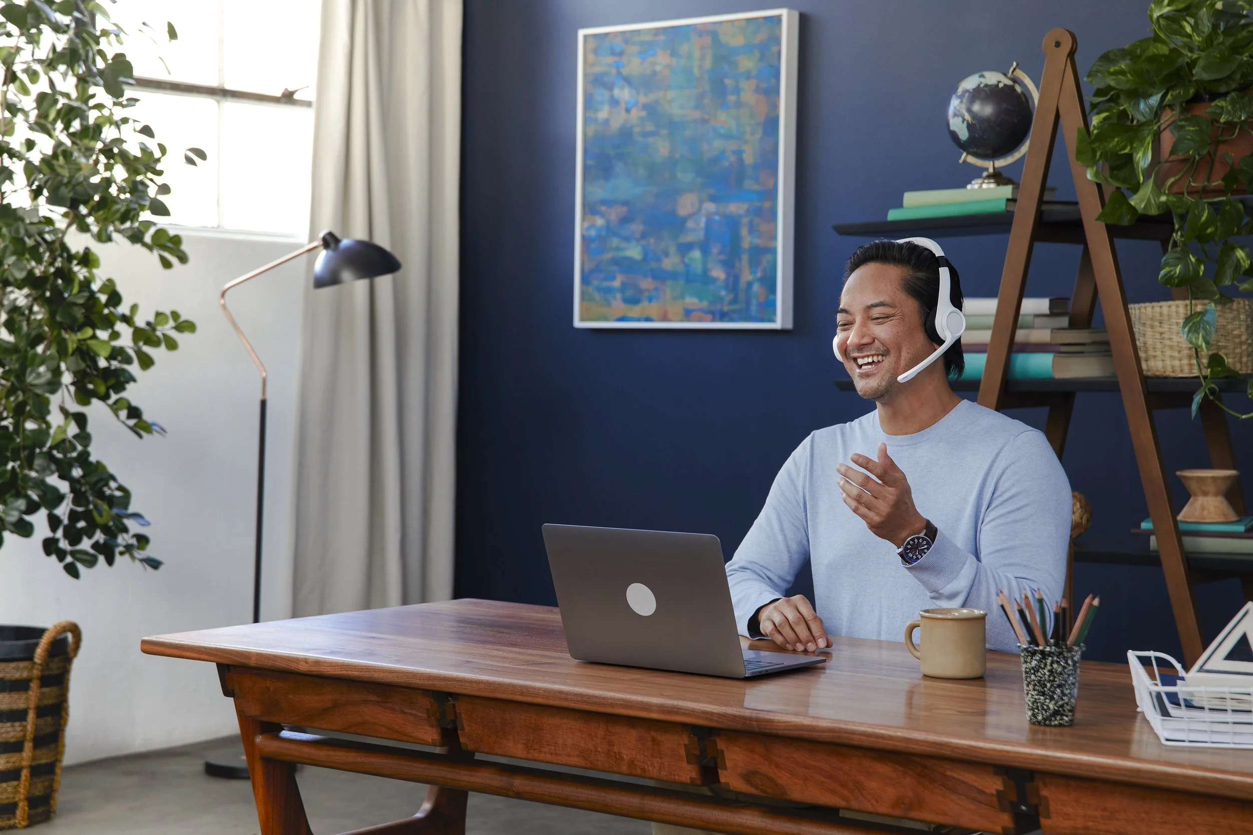A man sitting at a wooden desk with a laptop, wearing a headset, smiling and speaking, in a bright home office with a blue wall, large plant to the left, and a bookshelf with globe, books, and decor.