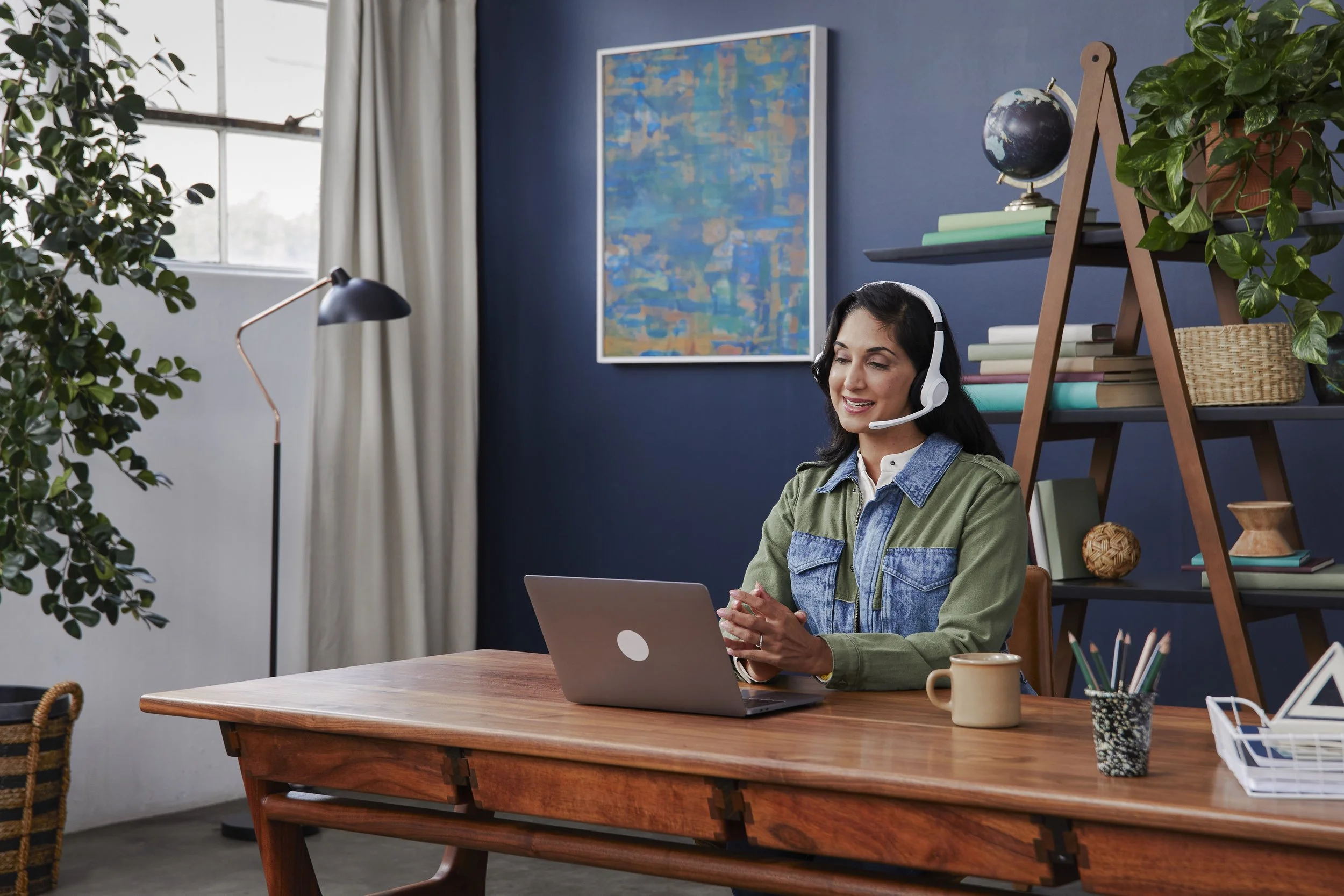 A woman wearing a headset sits at a wooden desk using a laptop in a home office decorated with plants, books, artwork, and a globe.