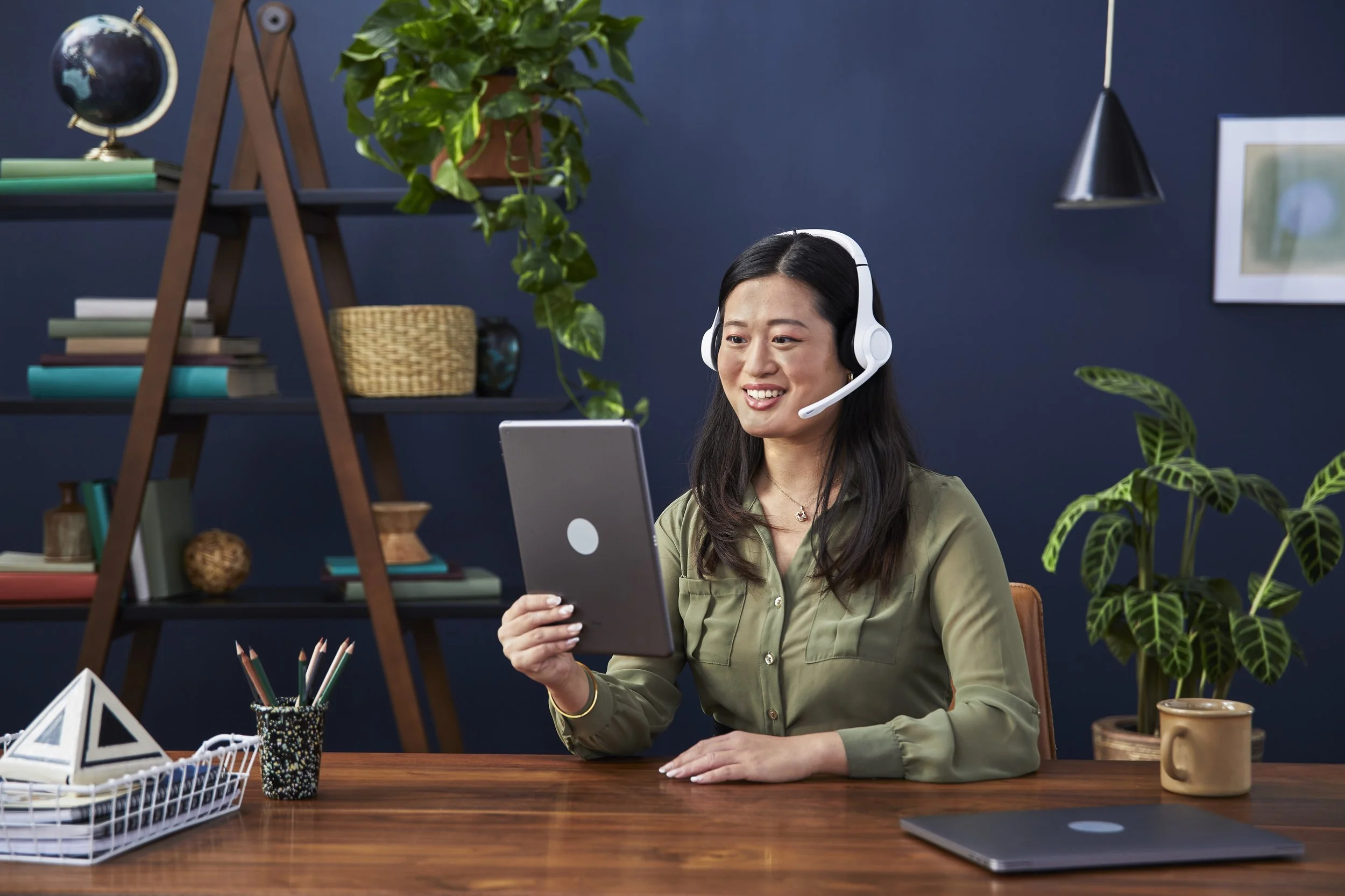 A woman with long dark hair wearing a headset and a green blouse sitting at a wooden desk with a tablet in her hand, smiling. The background features a blue wall with a shelf holding books, a globe, and potted plants, along with a framed picture and 