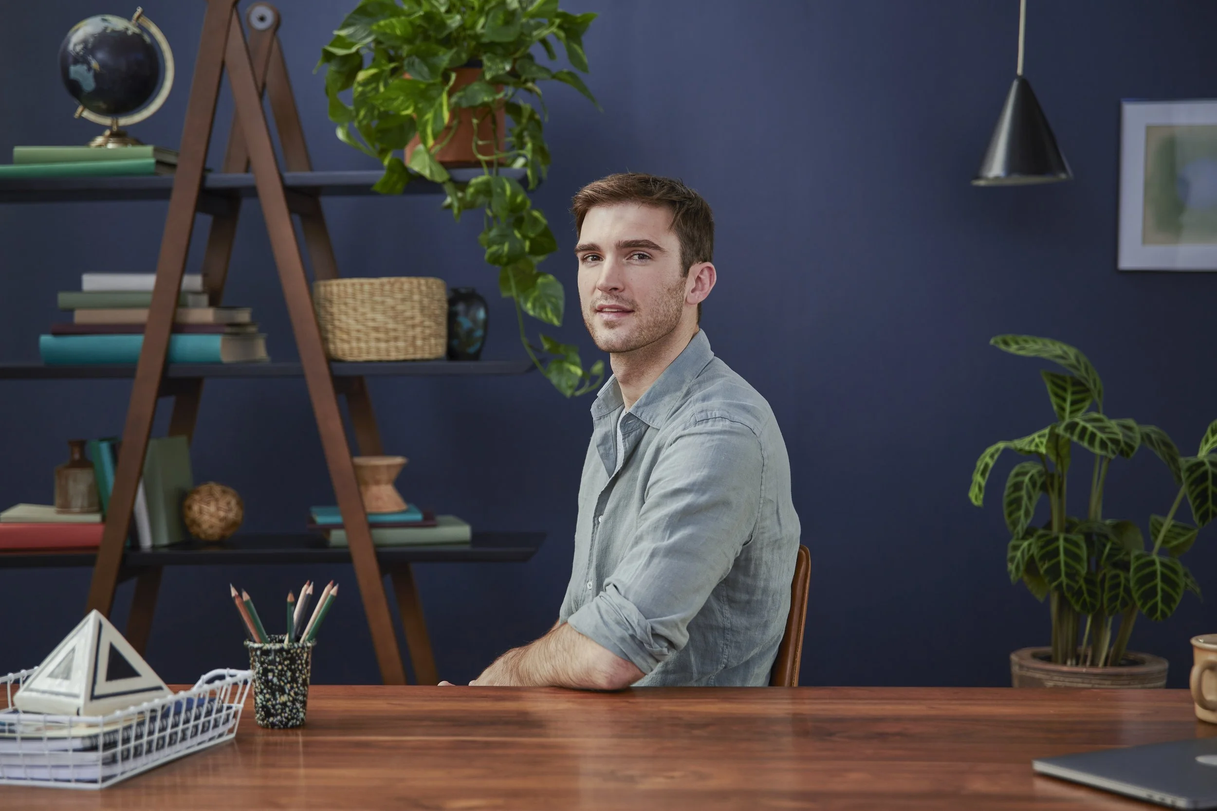 A young man with short brown hair and a light blue shirt sits at a wooden desk with various office supplies, including a geometric paperweight, a jar of colored pencils, and a white wire basket. Behind him is a dark blue wall with a bookshelf holding
