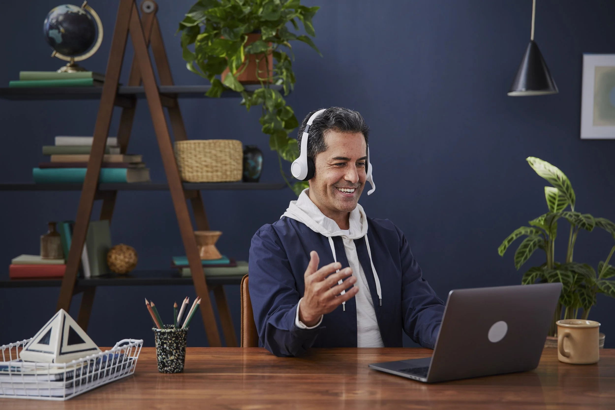 A man wearing a headset and a hoodie is sitting at a wooden desk with a laptop, smiling and gesturing with his right hand. The background has a dark blue wall with shelves holding books, plants, and decorative items, and there is a large leafy plant 