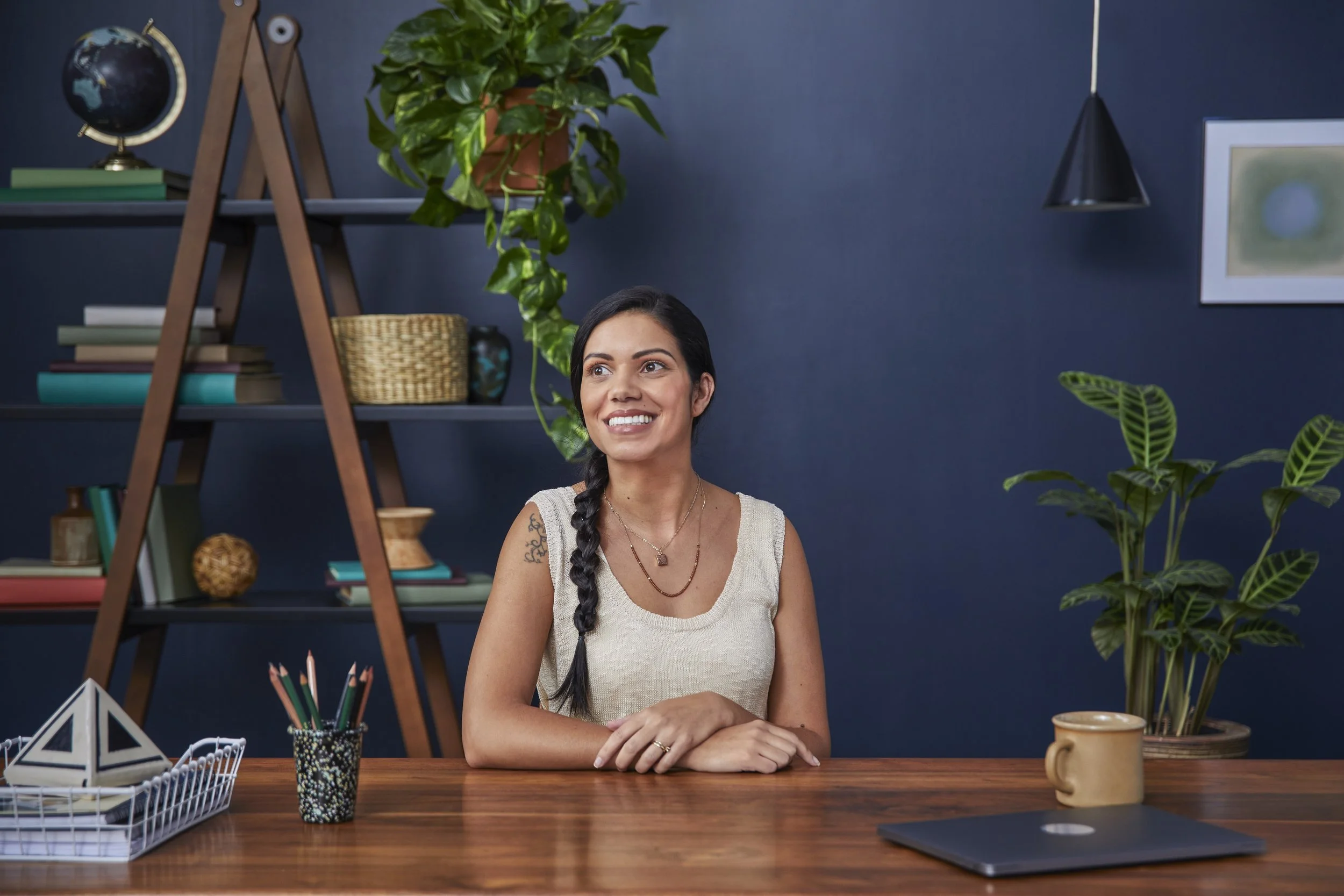 A woman with long dark hair in a braid smiling while sitting at a desk in a room with dark blue walls, decorated with shelves containing books, plants, and decorative items.