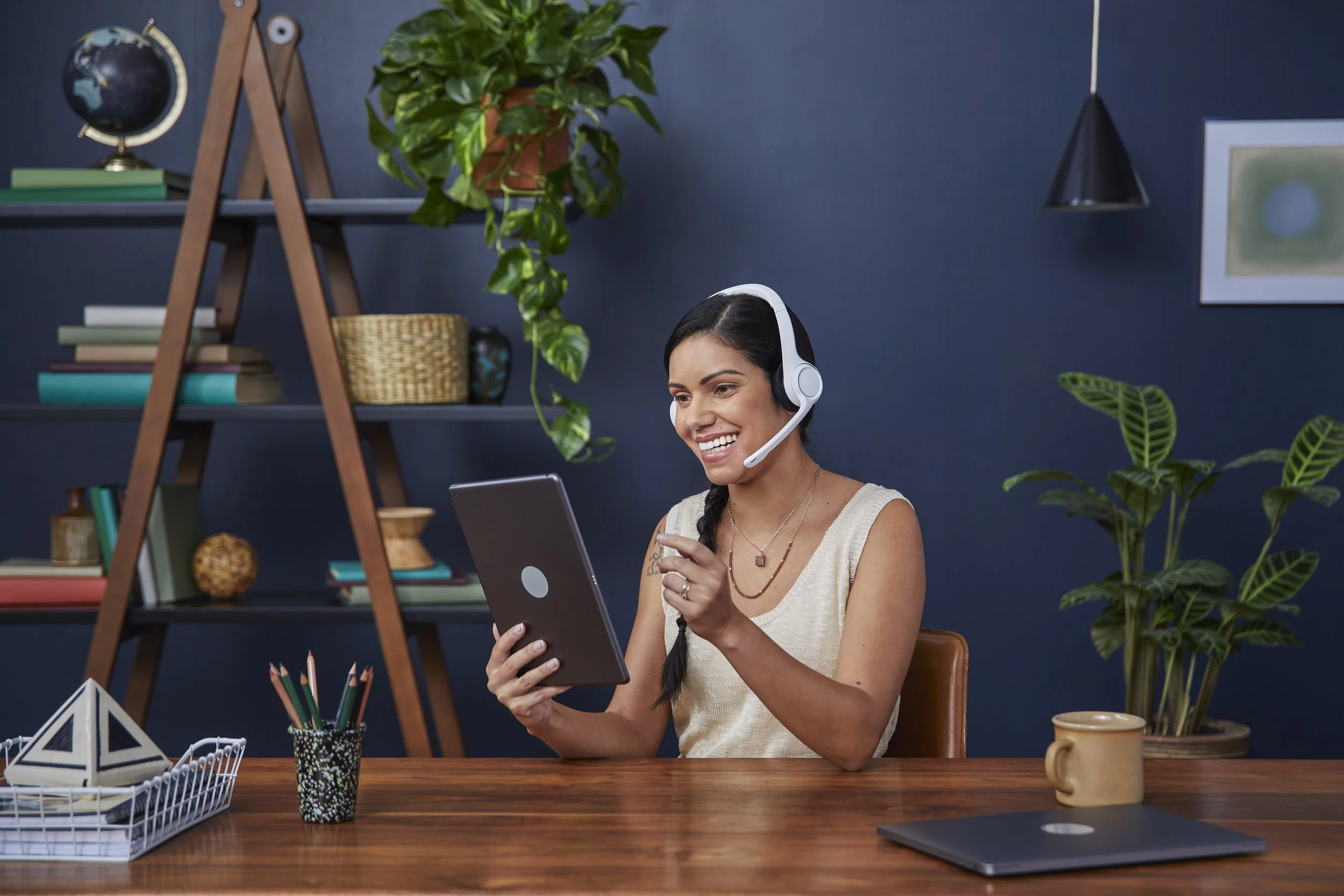 A woman smiling and wearing a headset is sitting at a wooden desk, holding a tablet, in a room decorated with plants, books, and office supplies.