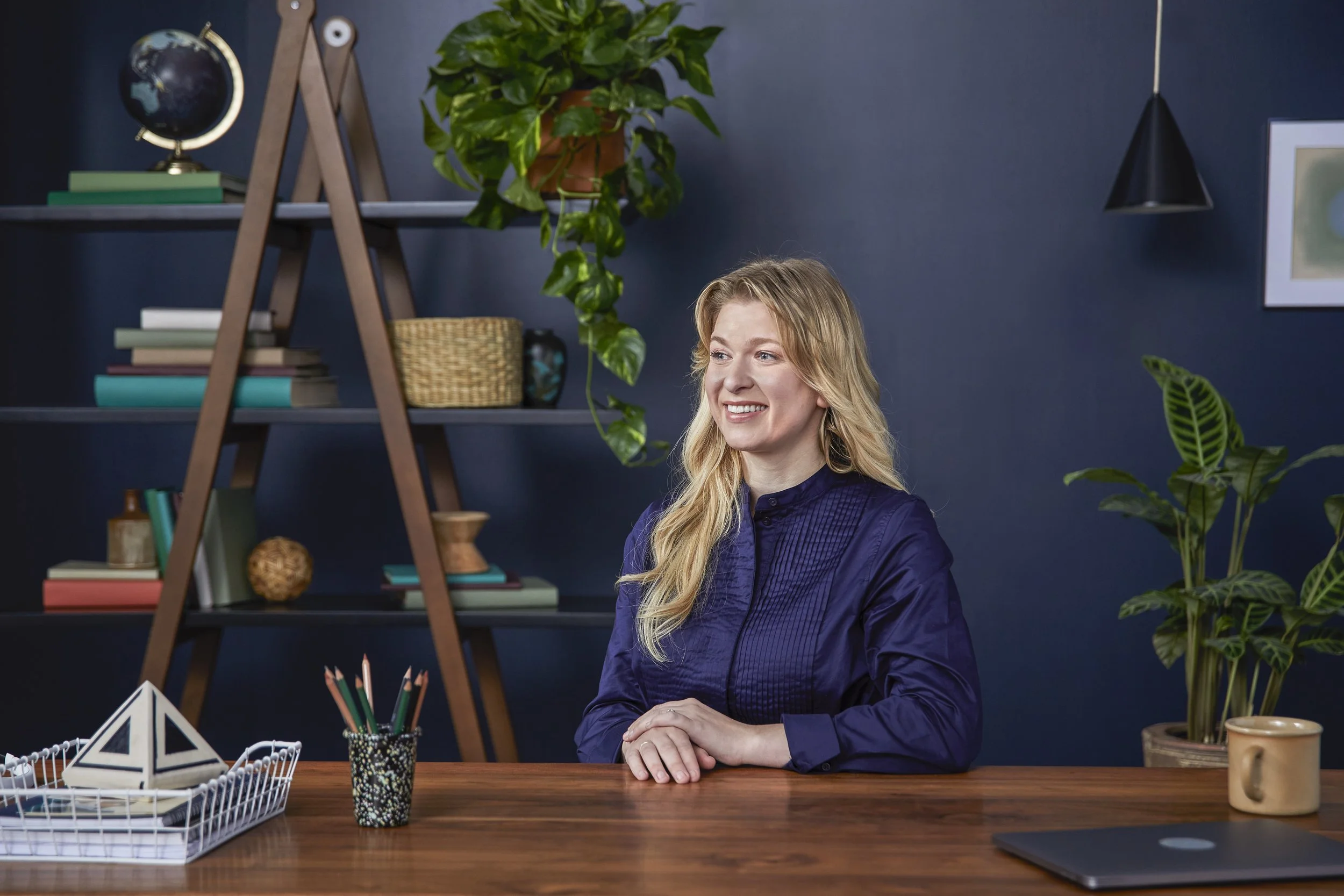 A woman with blonde hair smiling and sitting at a wooden desk with her hands clasped. The desk has a decorative cup with pencils, a geometric paperweight, a white basket, and a closed laptop. Behind her is a dark blue wall with a shelf holding books,