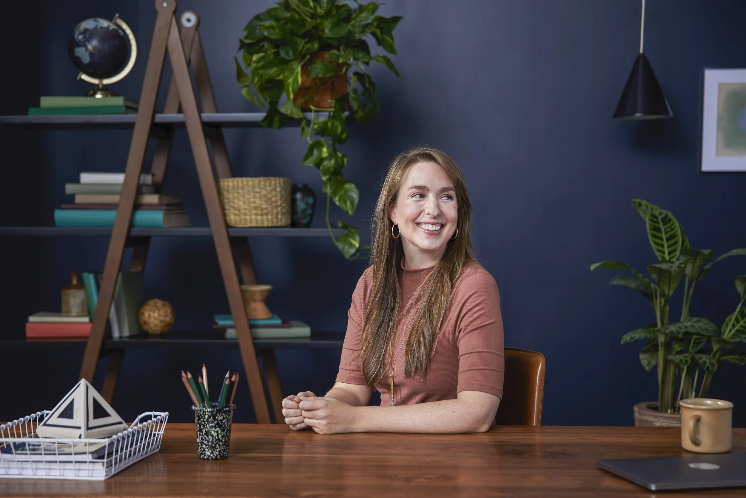 A woman with long light brown hair, wearing a pinkish tan top, is smiling and sitting at a wooden desk in front of a dark blue wall. Behind her is a black shelf with books, a globe, a plant, and decorative items. There are various plants in the scene