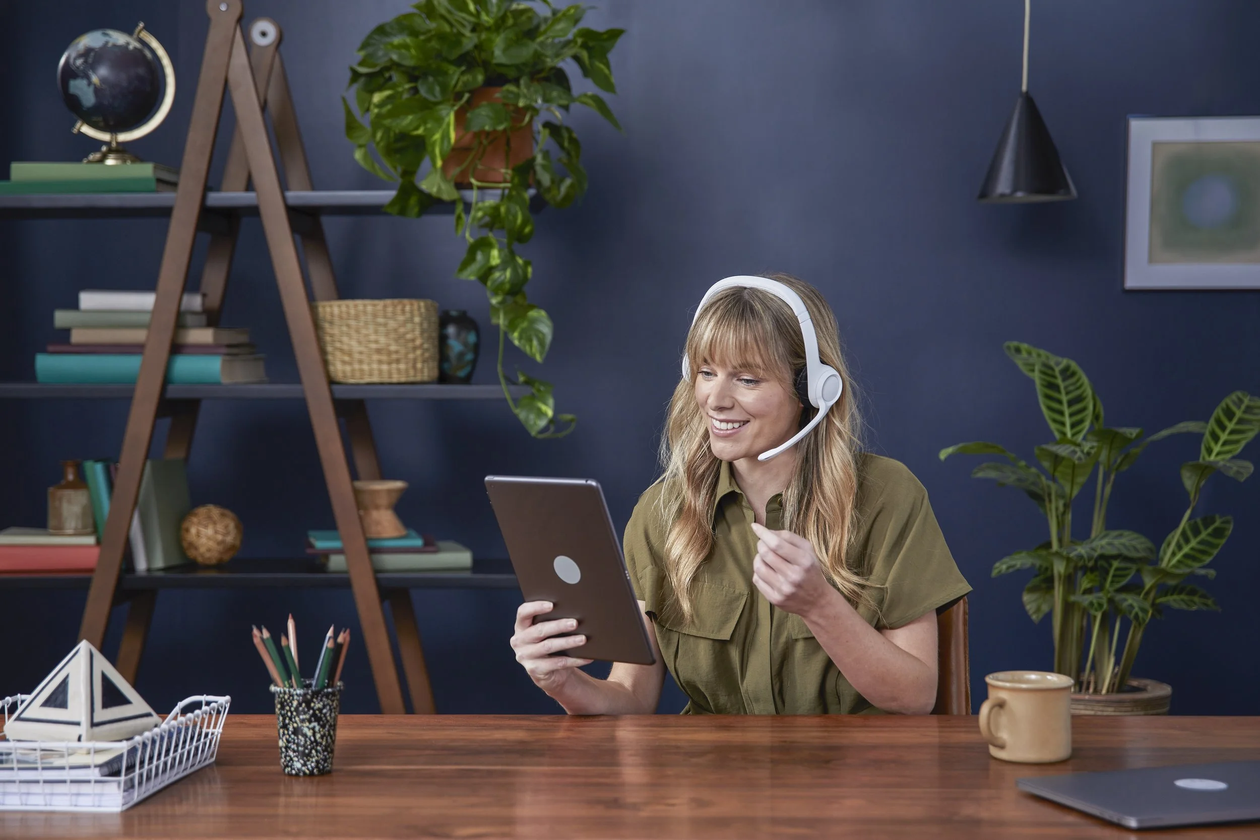 A woman with long blonde hair wearing a khaki shirt and white headset sitting at a wooden desk, holding a tablet and looking at it smiling. The background has a dark blue wall with a ladder-style shelf with books, a globe, houseplants, and a hanging 