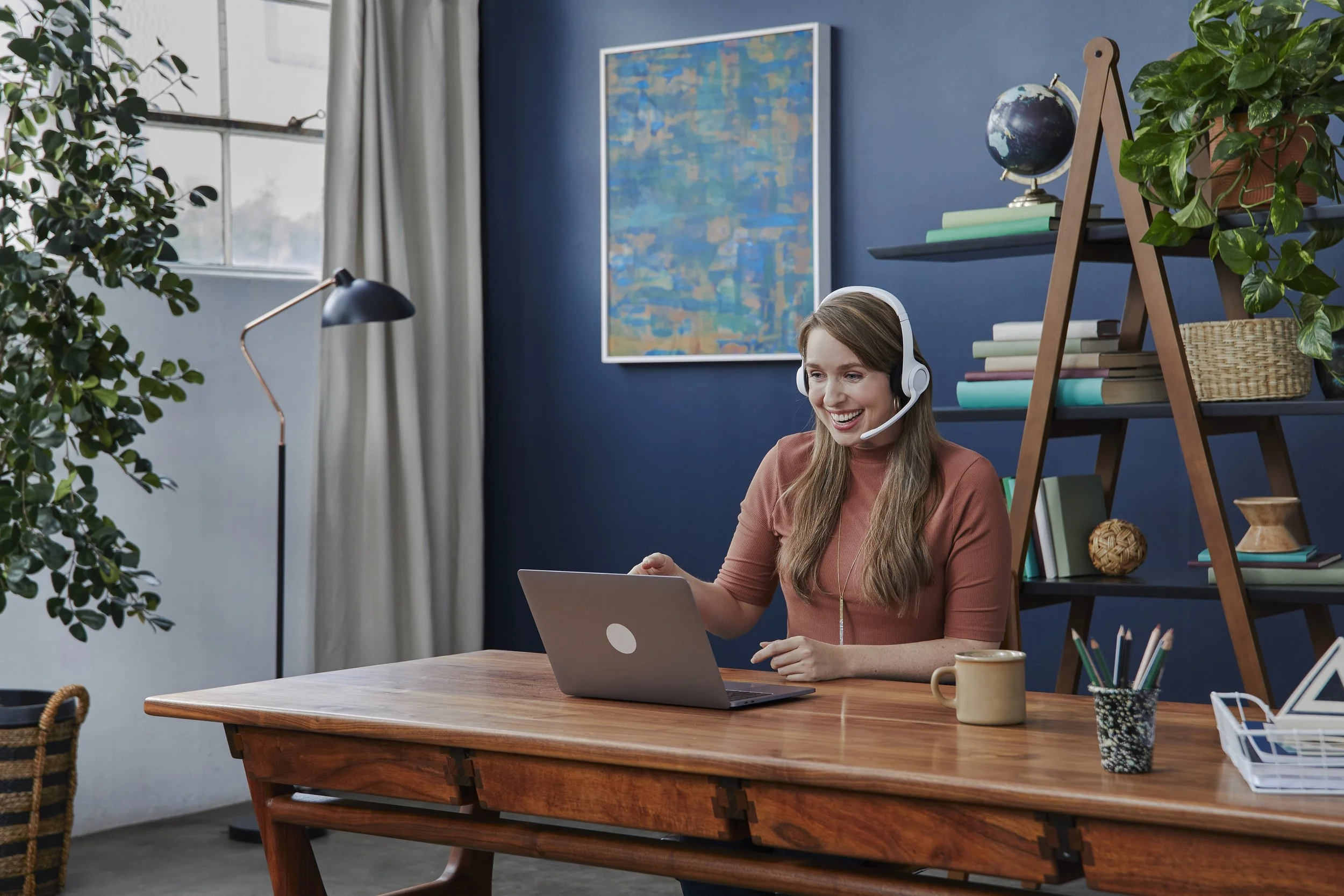 A woman sitting at a wooden desk in a home office, wearing a headset and smiling while looking at a laptop. The room has a large window, a blue painted wall with abstract art, a green plant, and a wooden bookshelf with various books, a globe, and dec