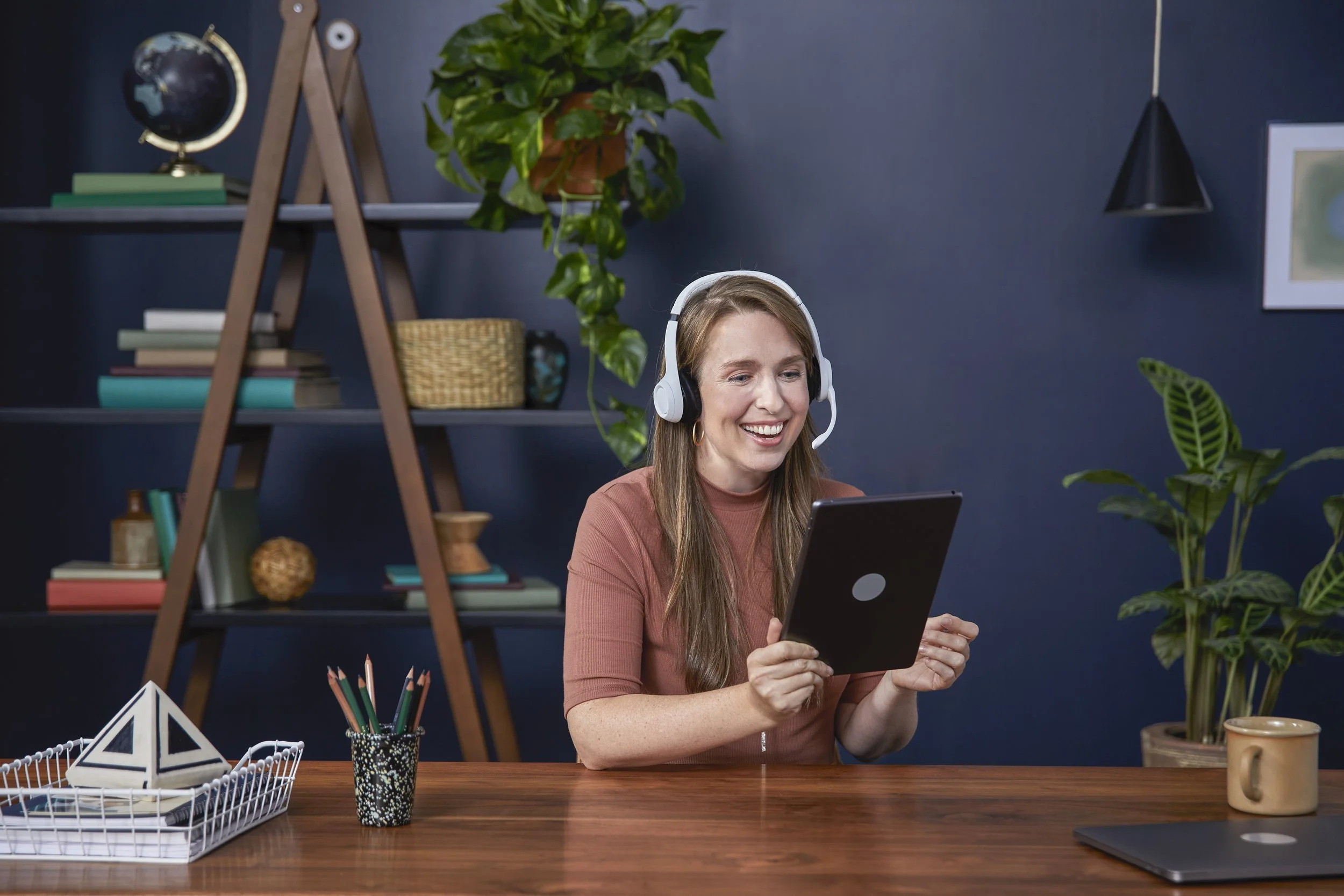 A woman with long hair and a warm-colored top sitting at a wooden desk, looking at a tablet device with a smile, wearing a white headset with a microphone.