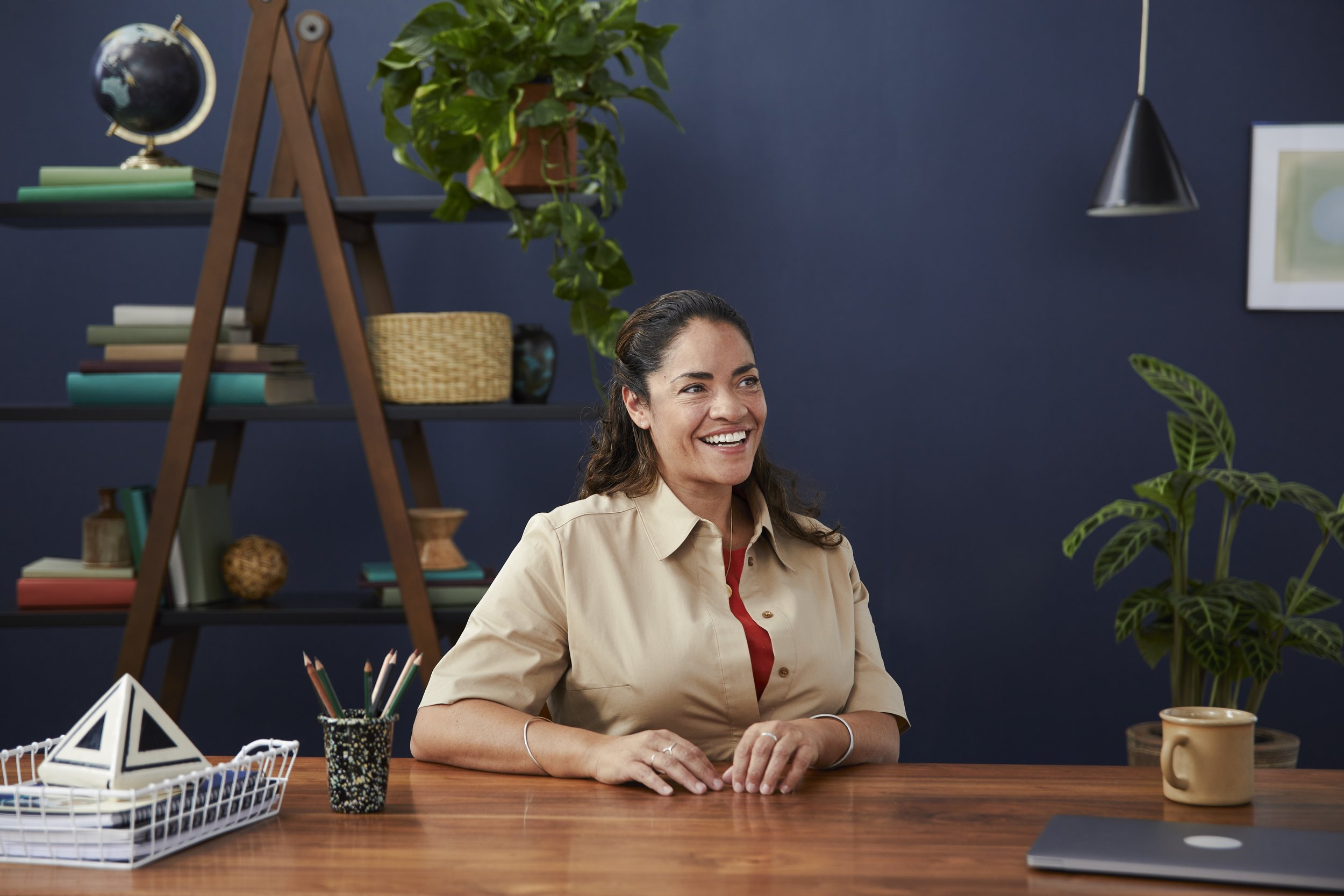 A woman sitting at a wooden desk in an office, smiling. Shelves with books, a globe, and decorative items are in the background, along with a plant and a framed picture on the wall.