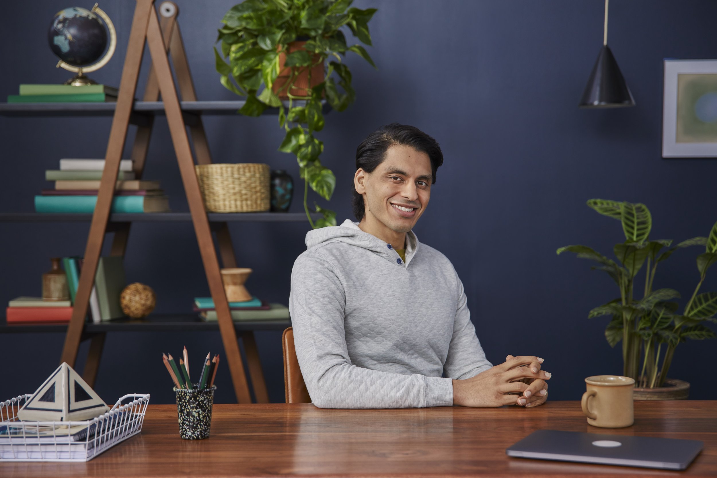 A young man with dark hair, wearing a light gray hoodie, sitting at a wooden desk in a room with dark blue walls, smiling and holding a mug. There are bookshelves with colorful books, plants, a globe, and decorative items in the background, with pens