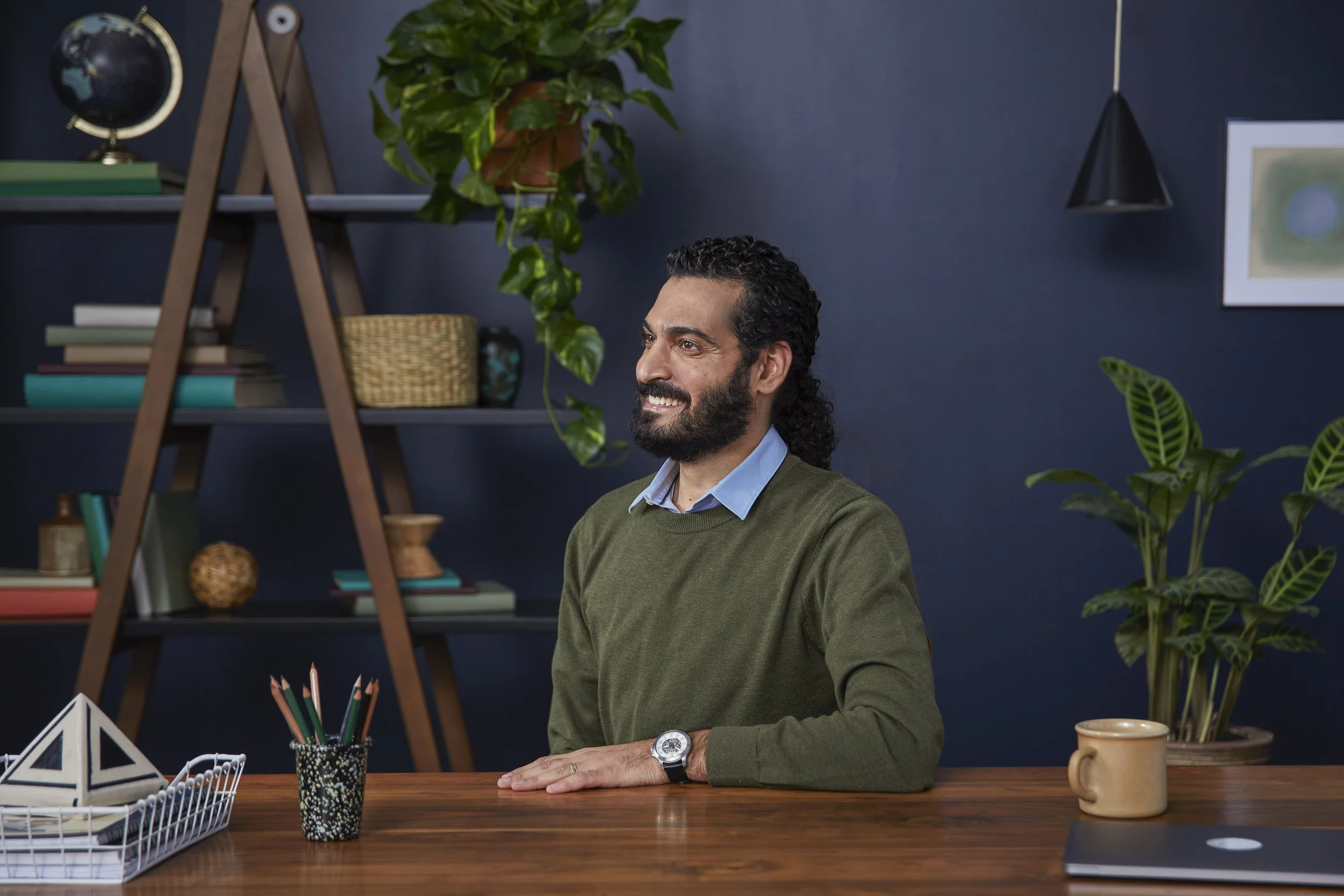 A smiling man with dark curly hair and a beard sitting at a wooden desk in a room with blue walls, surrounded by houseplants, books, and office supplies.