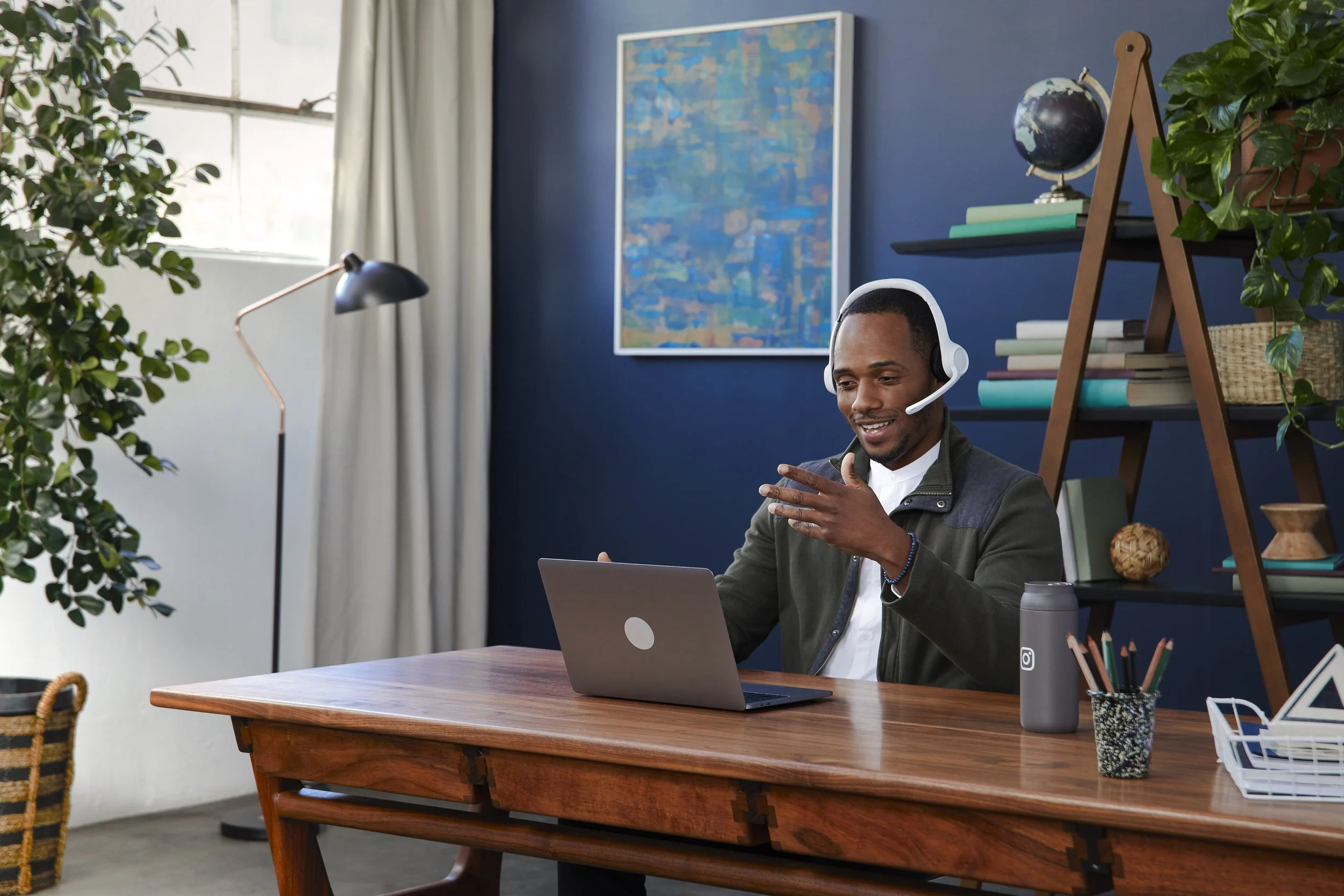 A man sitting at a wooden desk using a laptop while wearing a headset, in a modern, colorful home office with plants, books, and artwork on the walls.
