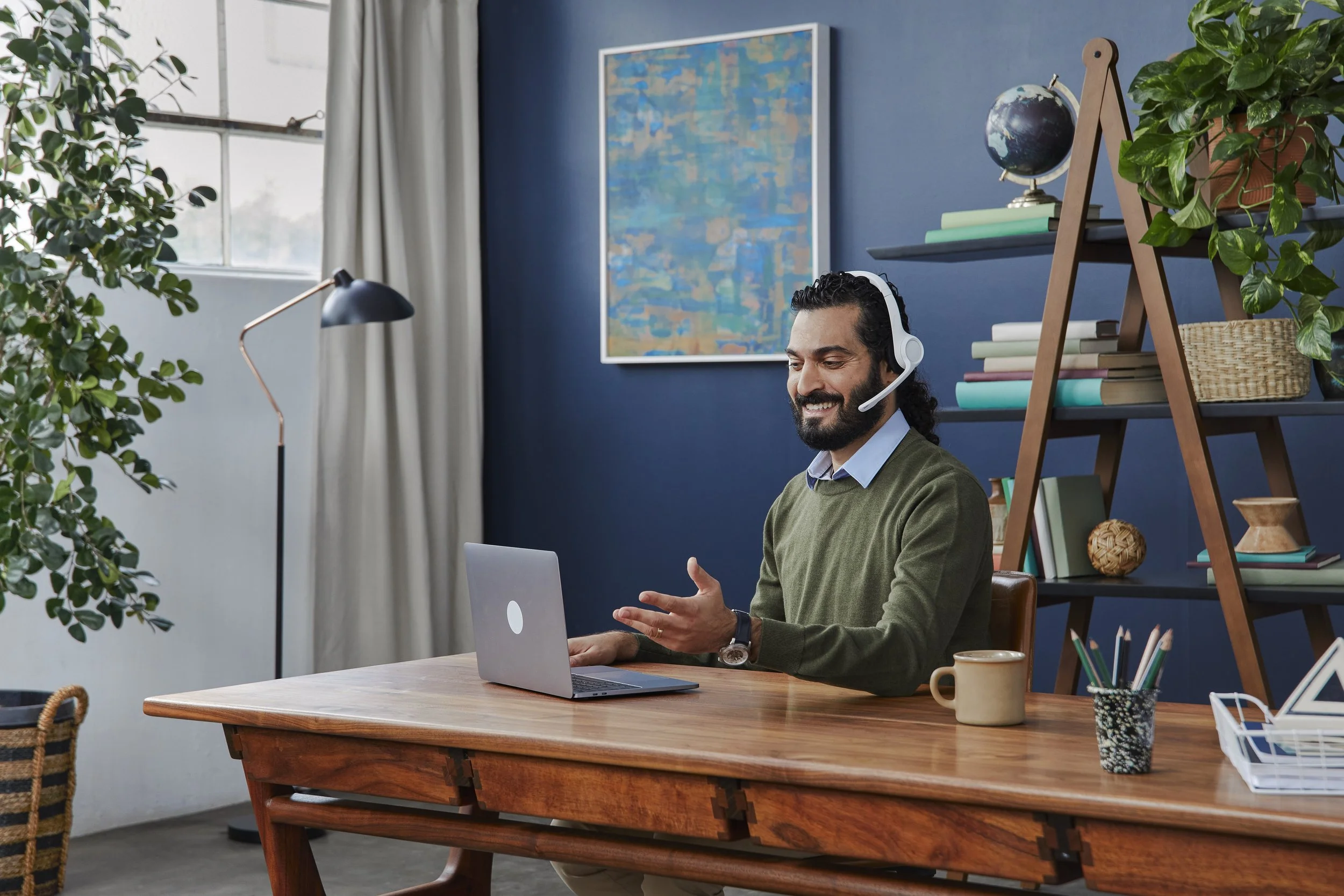 A man with long curly hair and a beard, wearing a headset and a green sweater, sits at a wooden desk using a laptop in a home office with plants, books, a globe, and a large abstract painting on the blue wall.
