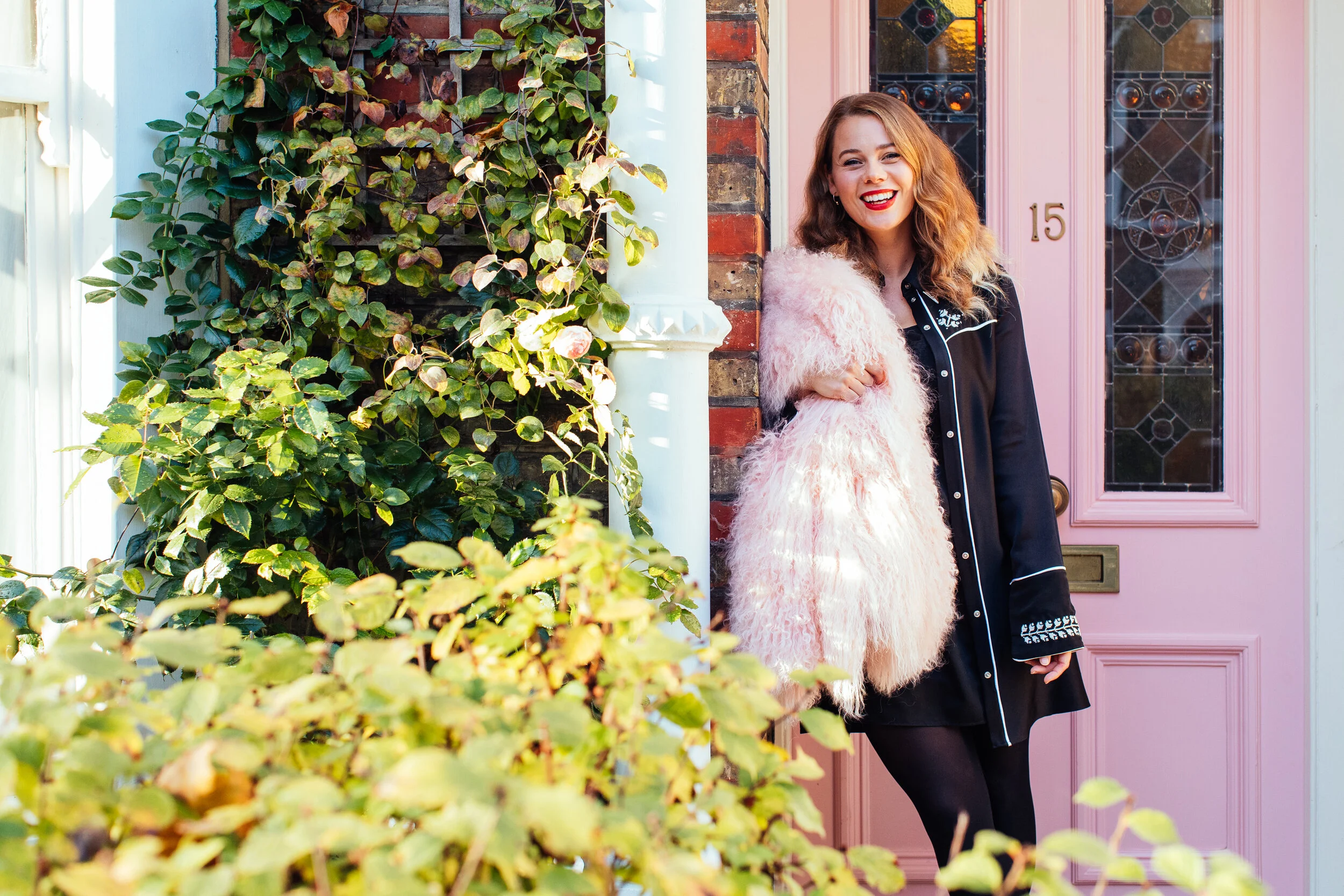 A woman with red curly hair, red lipstick, and hoop earrings, smiling, standing at a pink front door with house number 15, next to a brick wall and green leafy bushes.