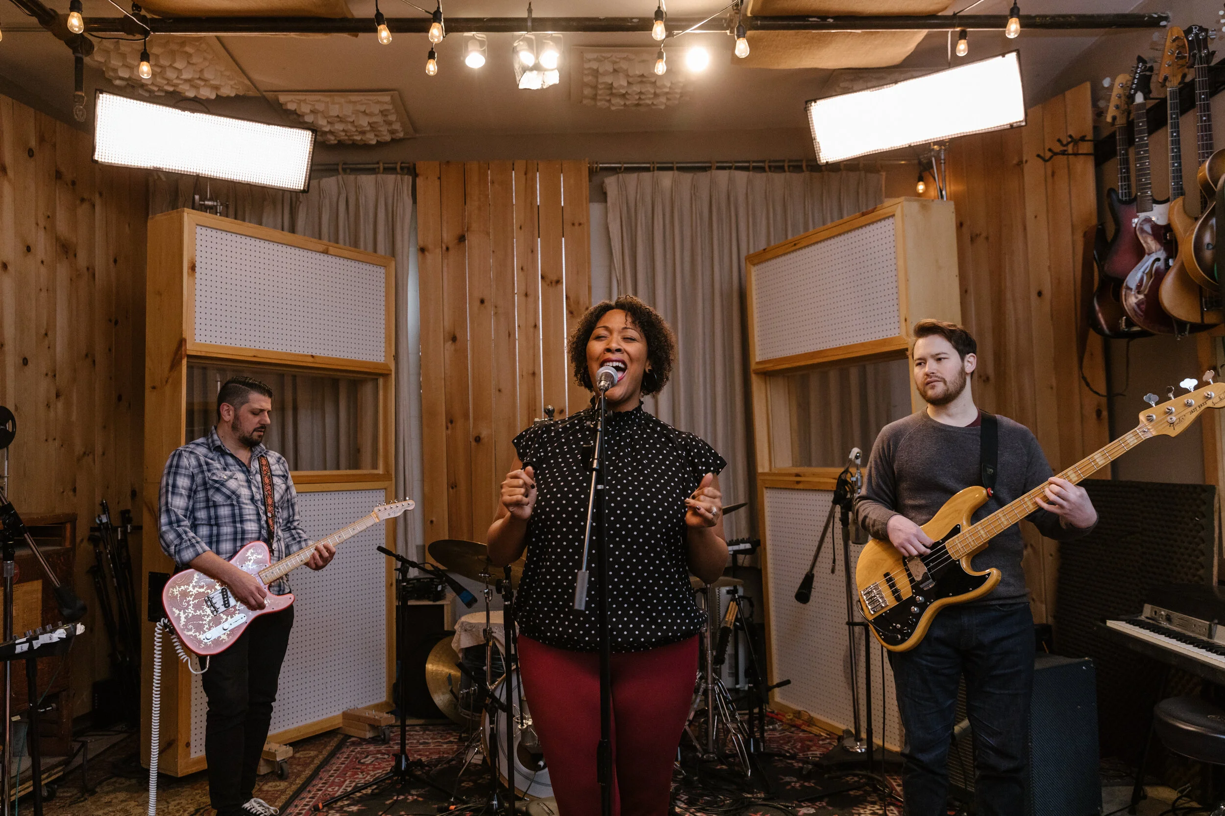 A band performs in a recording studio with wooden walls. The female lead singer is singing into a microphone, flanked by two male guitarists, one on electric guitar and the other on bass, with drums and a keyboard in the background. Overhead, string lights hang from the ceiling, creating a cozy atmosphere.