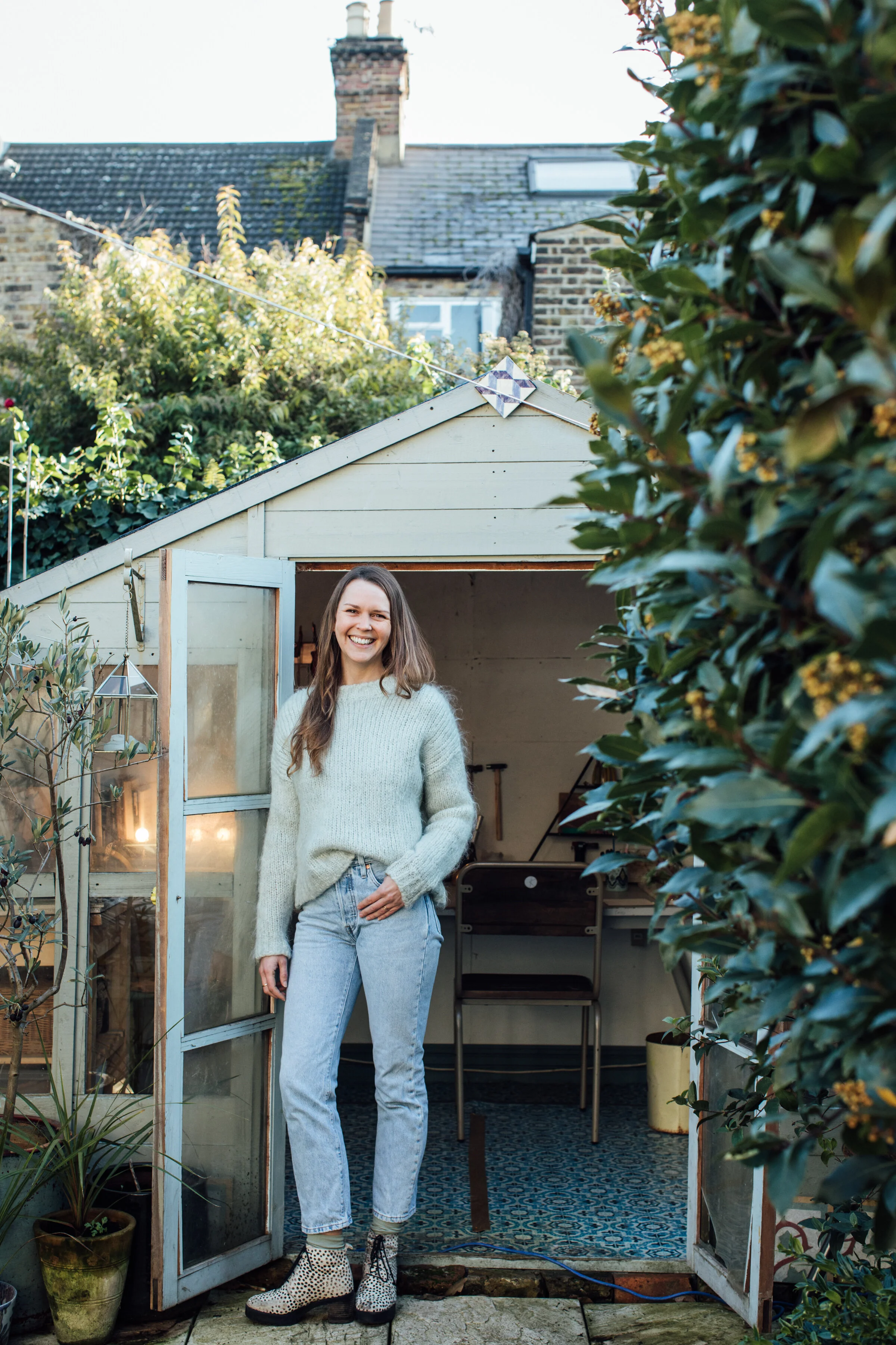 A young woman with long brown hair, wearing a light gray sweater, light blue jeans, and leopard-print boots, stands in a garden shed doorway smiling. The shed has glass windows and is surrounded by green plants and bushes. The background shows a bric