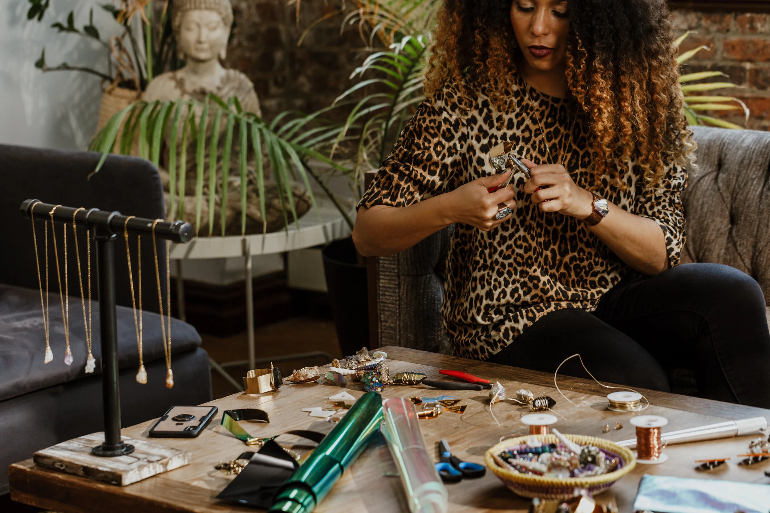 Woman with curly hair wearing a leopard print top sitting at a table working on jewelry making, surrounded by tools, jewelry pieces, and materials in a room with plants and a brick wall.