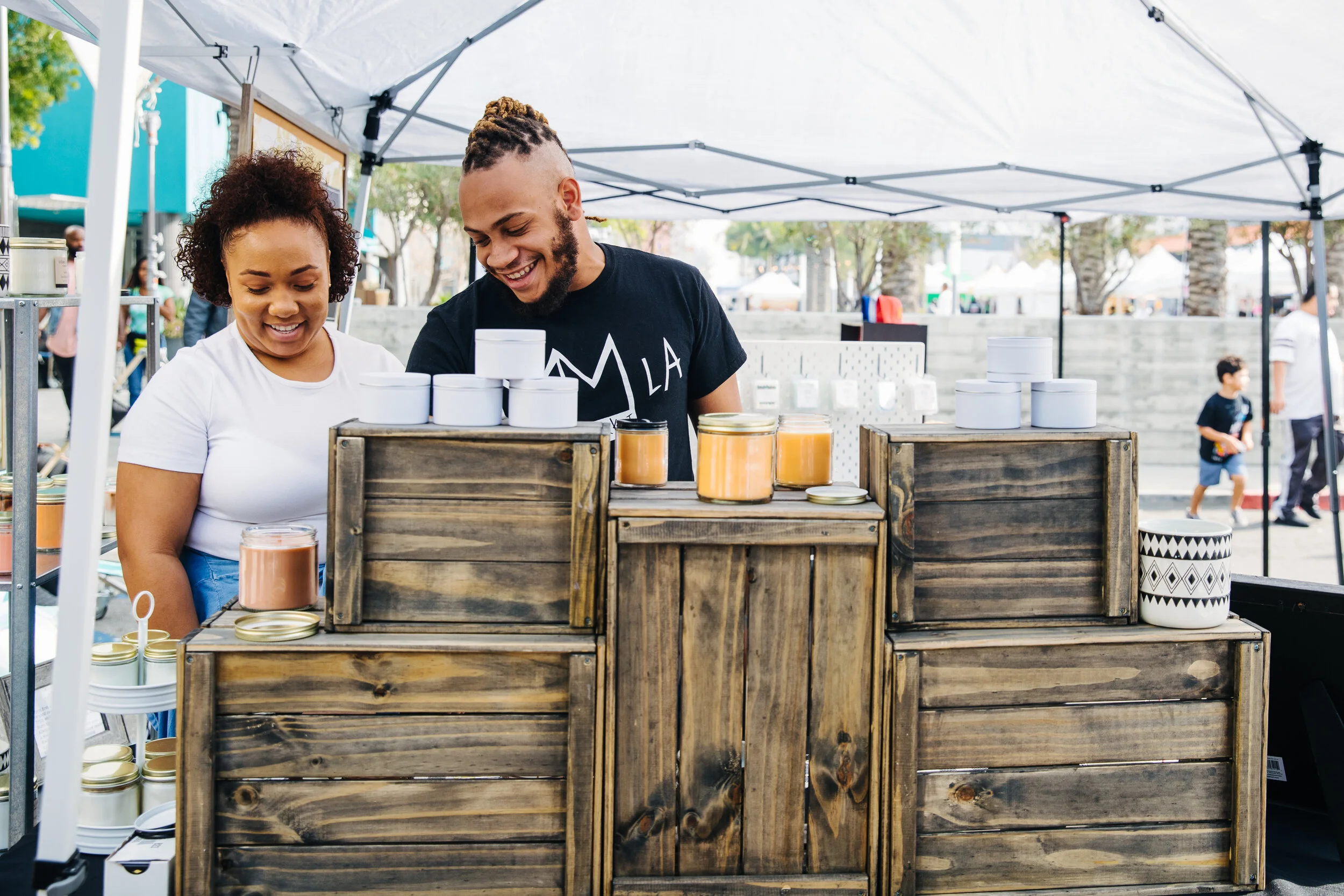Two people smiling at a market stall with jars of honey displayed in front of them under a white canopy, with other market booths and people in the background.