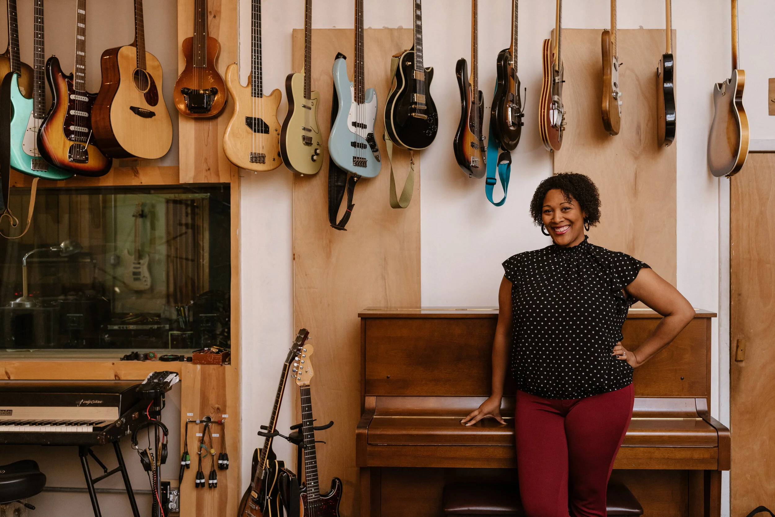 A woman with curly hair smiling in a room with a wall of various guitars and a keyboard. She is wearing a black polka dot blouse and red pants.