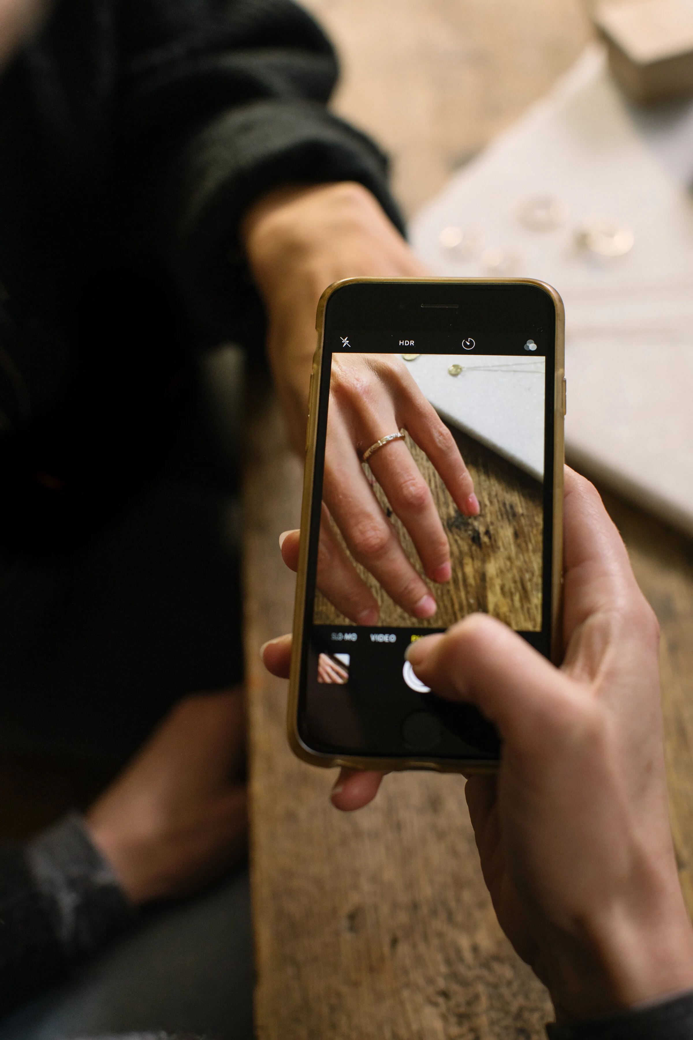 Person taking a photo of their hand and ring with a smartphone on a wooden table.
