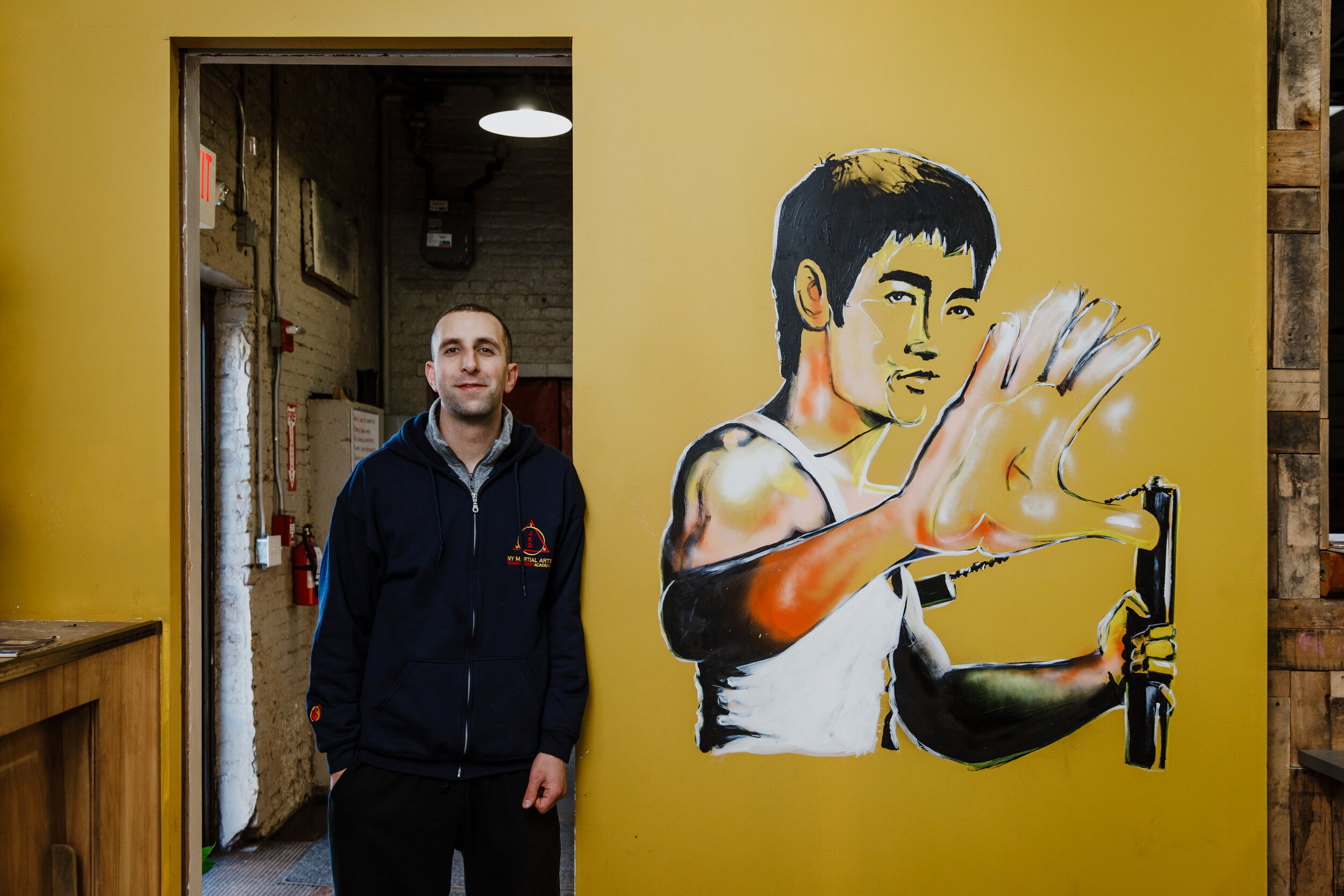 A man standing indoors next to a large mural of a female martial artist in a fighting stance with her hand up, painted on a yellow wall.