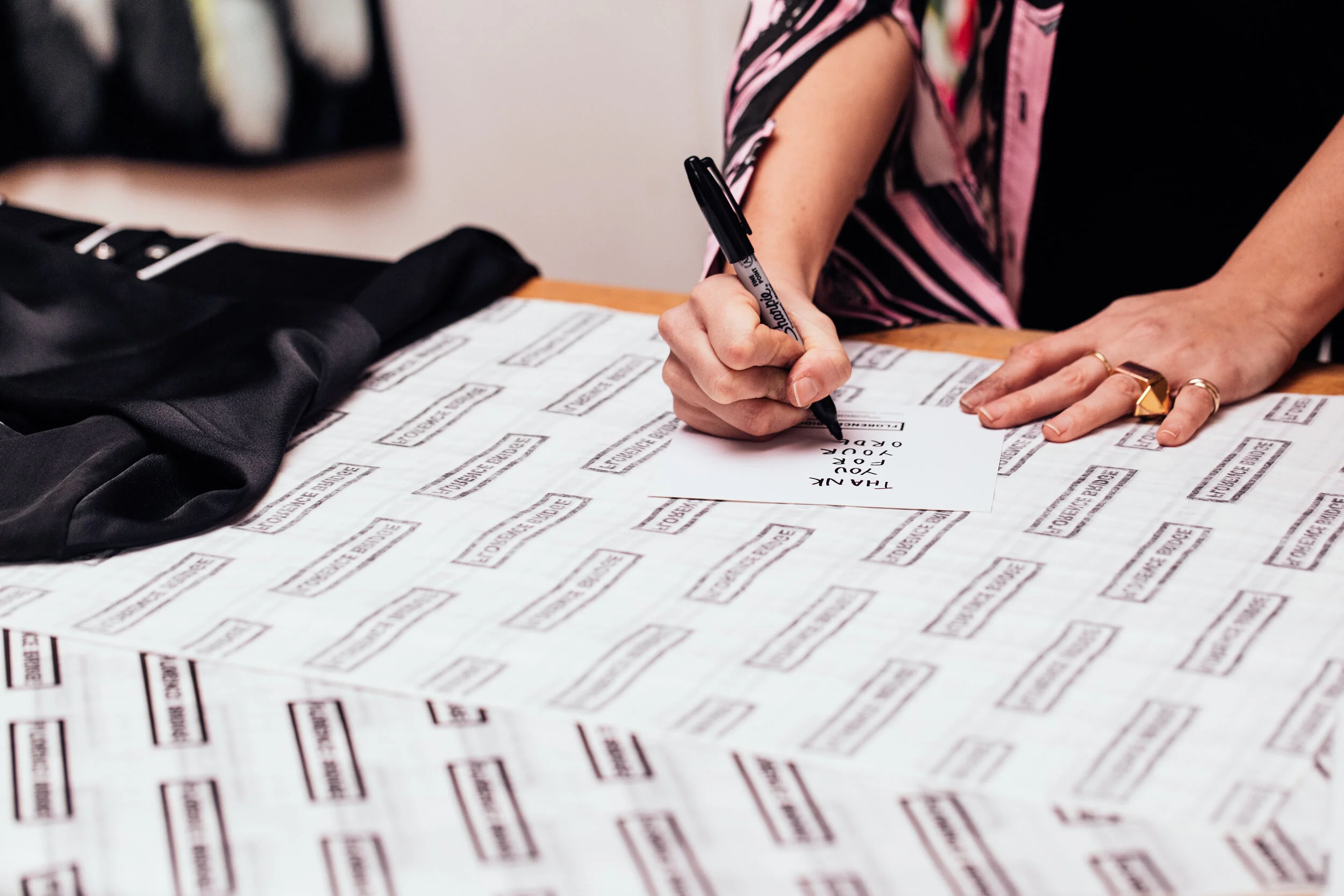 A person writing on a white paper with a black marker, surrounded by black and white printed paper that says 'TOURIST EXPERIENCE.' The person is wearing a gold ring and has a pink and black patterned shirt.