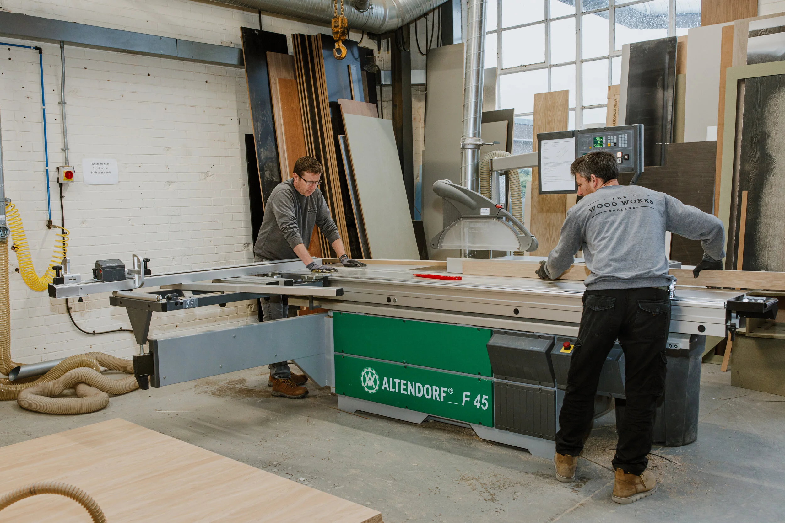 Two workers operating an Altendorf F 45 woodworking bandsaw in a woodworking shop, with various wooden panels stored vertically behind them.
