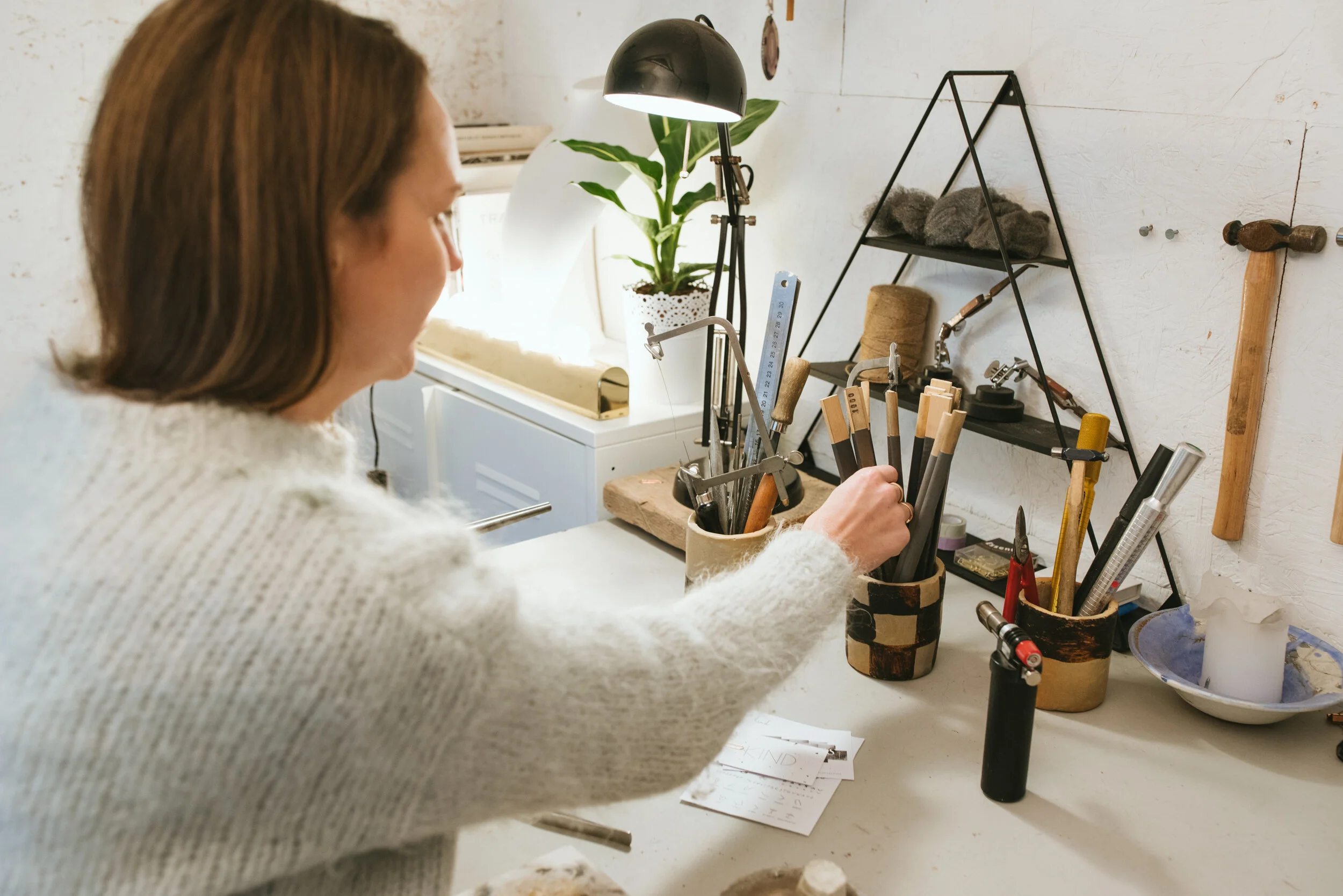 A woman working at a cluttered craft or jewelry making station with various tools, a plant, and notes on the table.