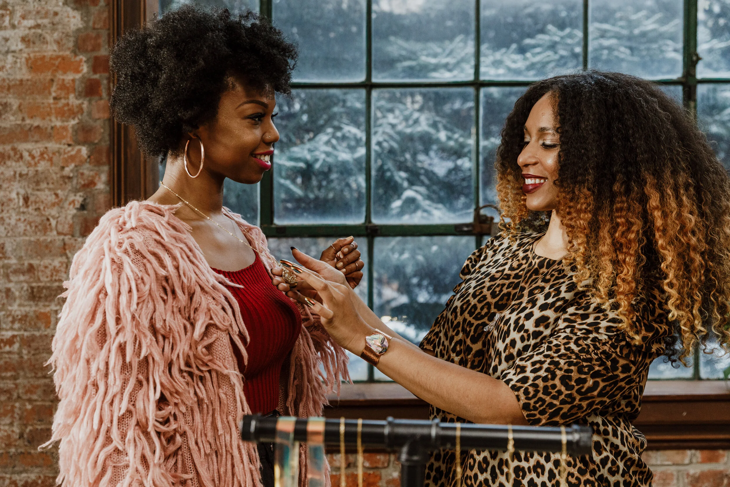 Two women facing each other smiling inside a brick-walled room with large windows showing snow outside. One woman has curly black hair, wearing large hoop earrings, a pink fuzzy cardigan, and a red top. The other woman has long curly brown hair, wearing a leopard print top, and is placing a ring on the first woman's finger.
