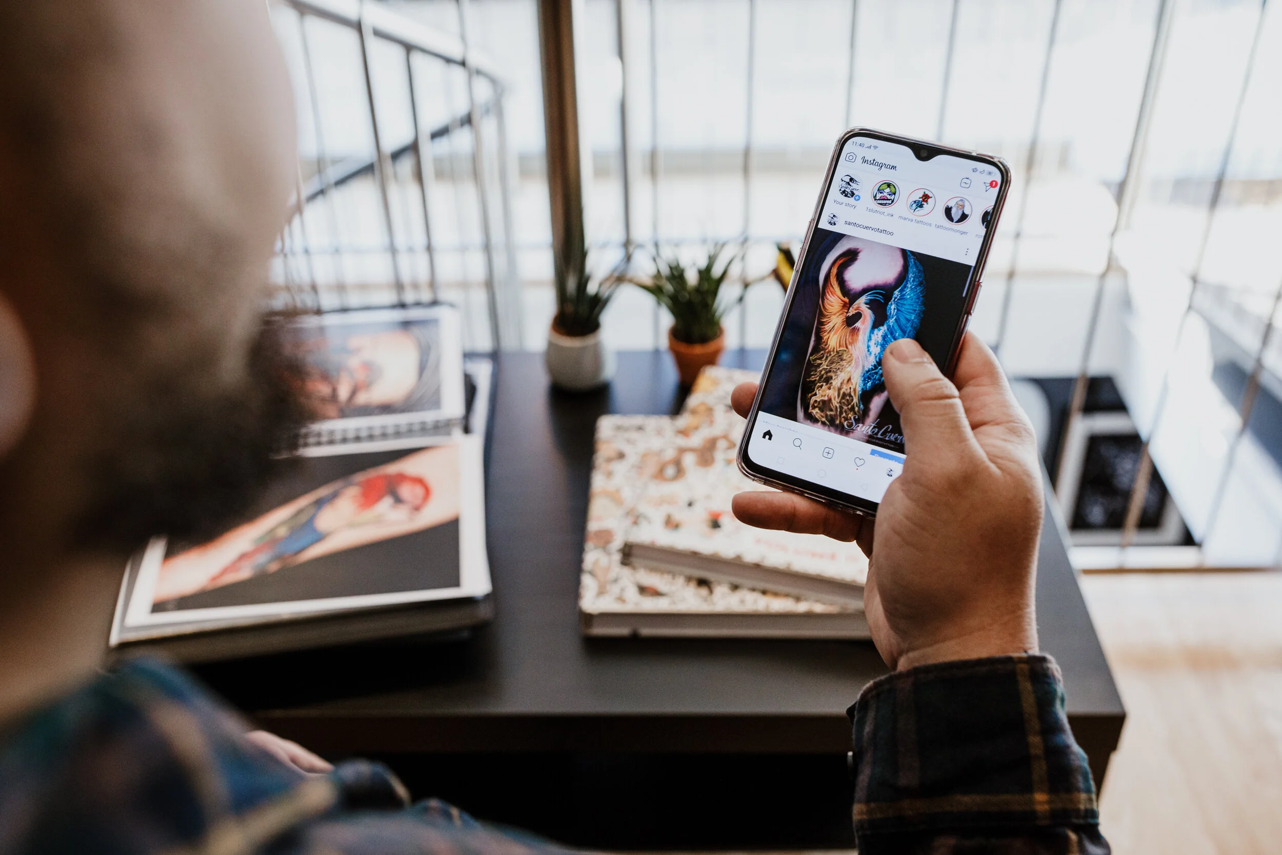 A person holding a smartphone displaying an Instagram page with a colorful lion artwork, surrounded by books and a potted plant on a table in a bright, modern space.