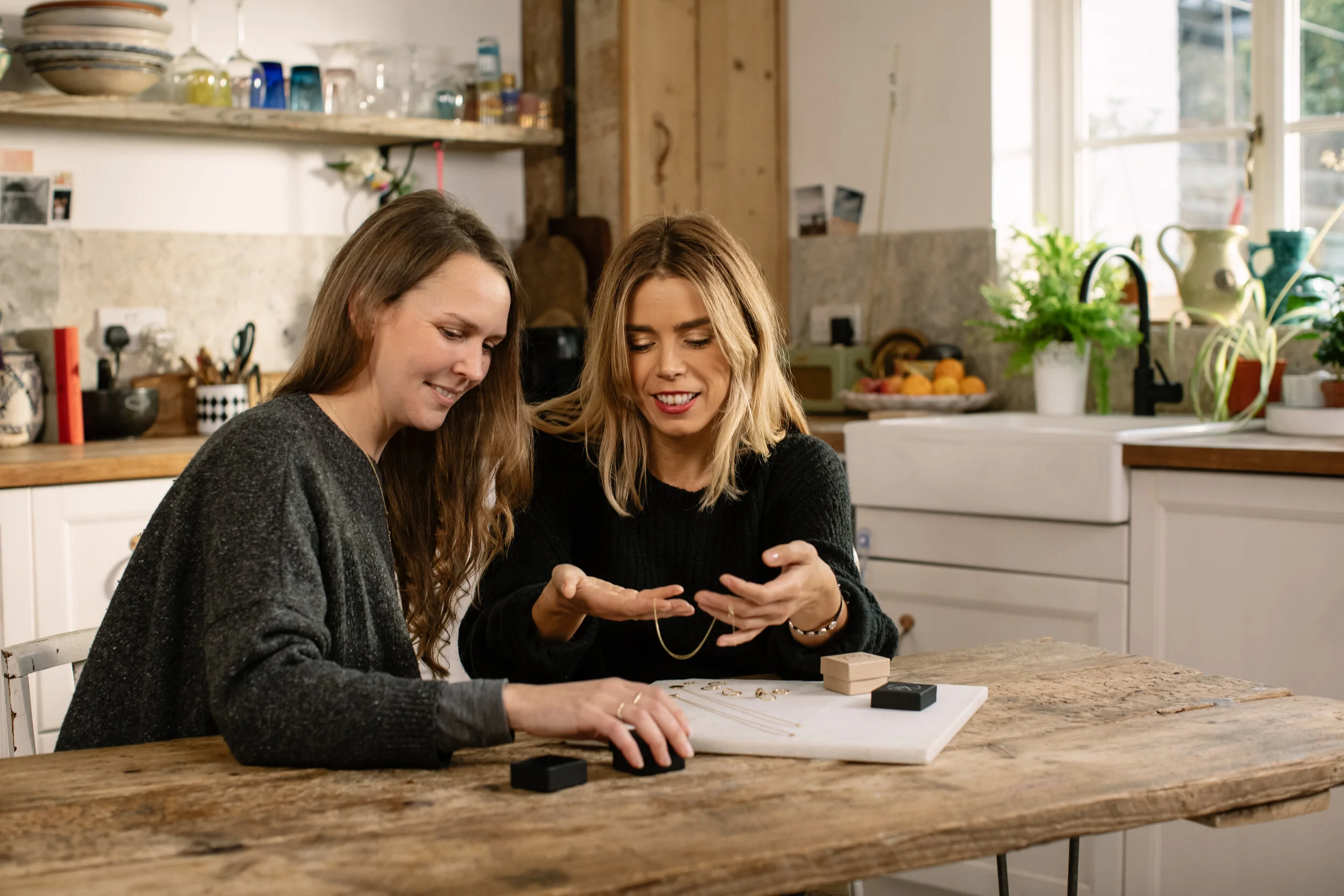 Two young women sit at a wooden kitchen island, smiling and looking at jewelry or small objects in one woman's hands.