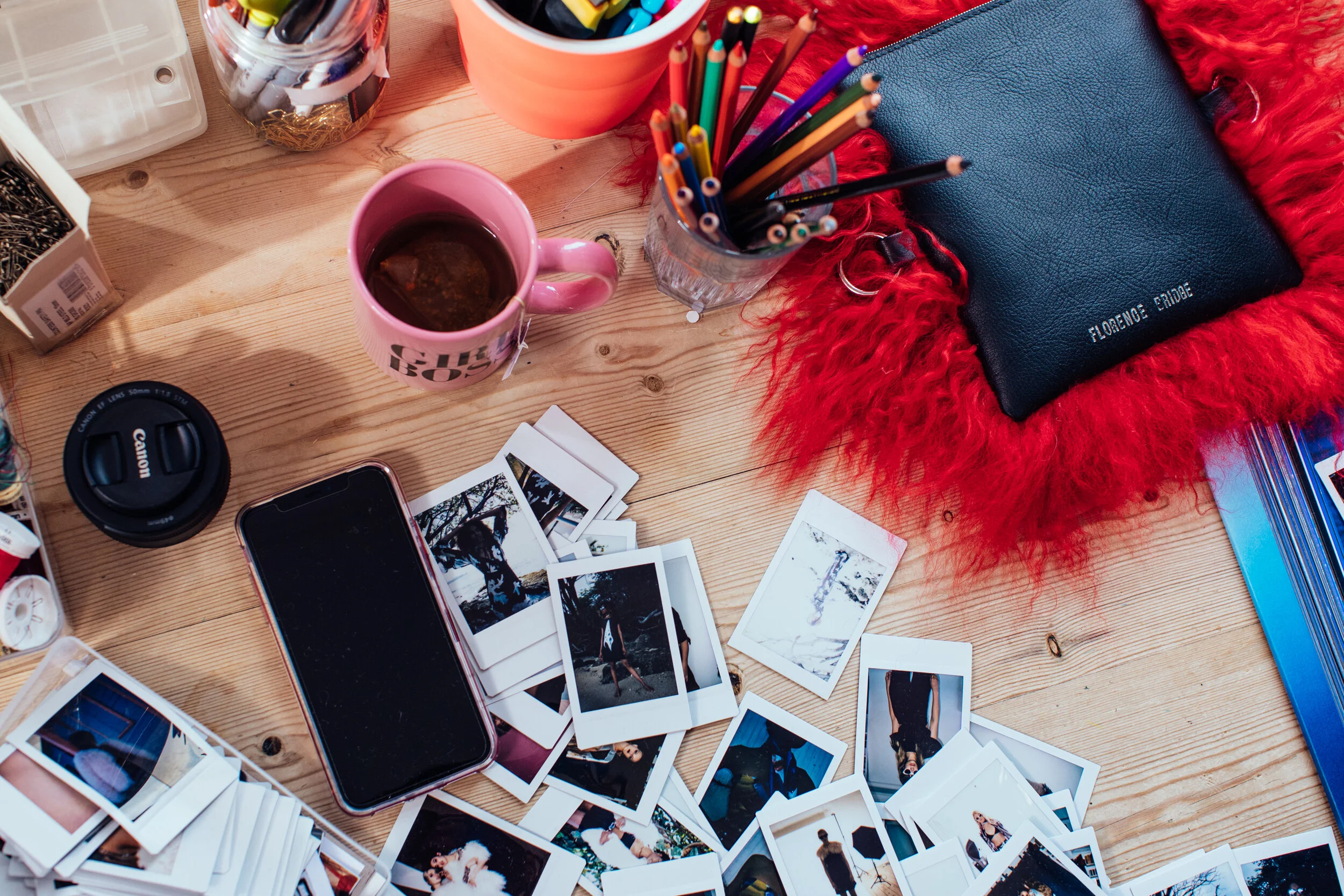 A wooden table with scattered instant photographs, a smartphone, a pink mug filled with tea, and various colorful stationery items including pencils and pens in glass containers. A closed blue wallet rests on a red furry cloth.