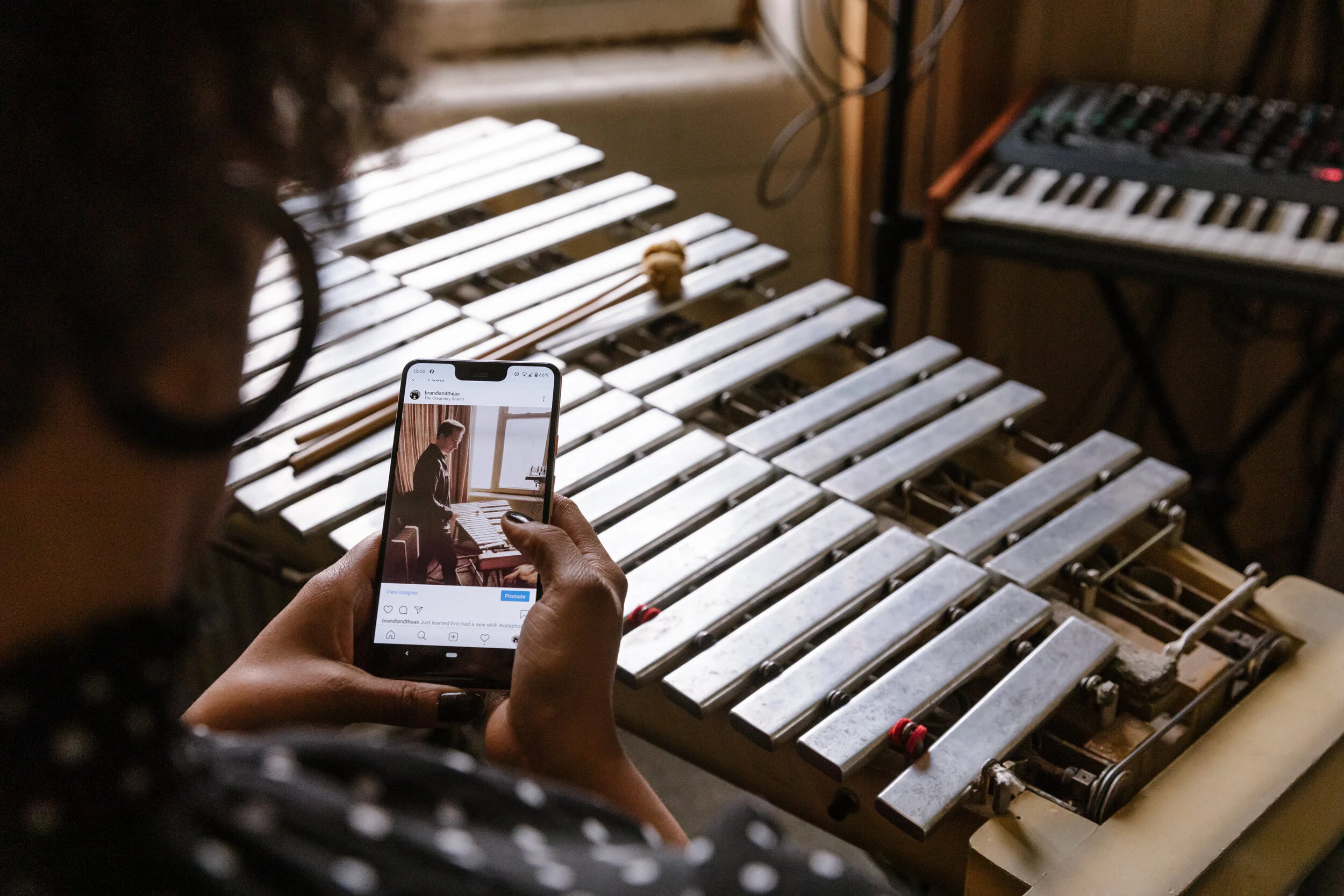 A person is taking a photo of a musical instrument, possibly a xylophone, with a smartphone. The instrument has metal bars and is in a room with a keyboard and some wiring.