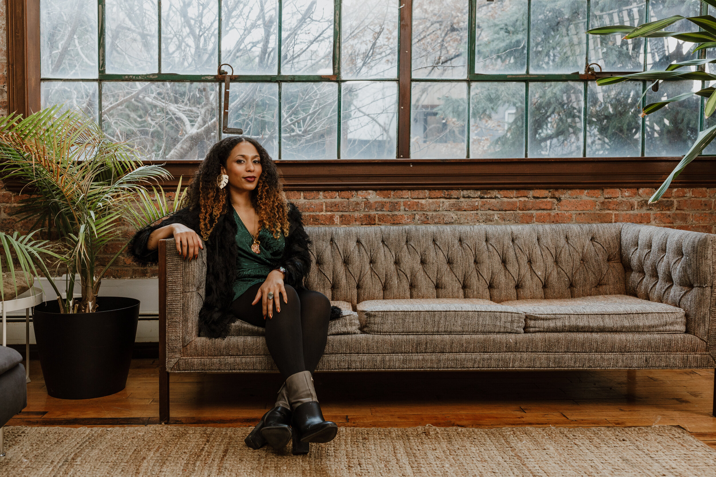 A woman sitting on a vintage tufted sofa in a cozy room with large industrial-style windows, wooden floors, and a brick wall, surrounded by plants.